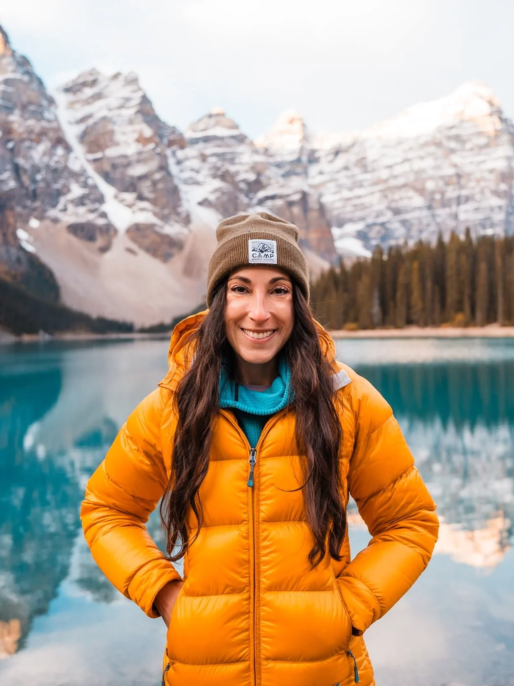 a woman in a yellow puffy jacket in front of a blue lake and snowy mountains