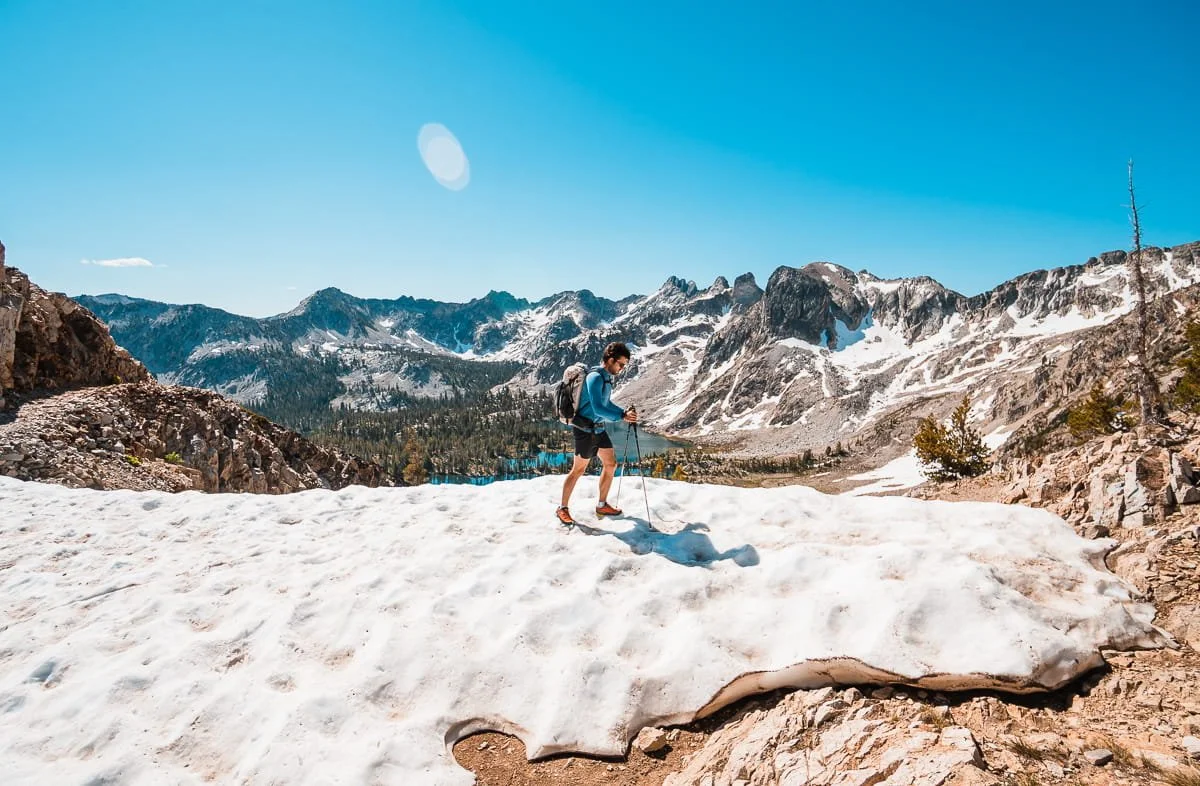 A man backpacking across a snowfield, wearing a blue sun hoodie, with rocky peaks and a lake in the background