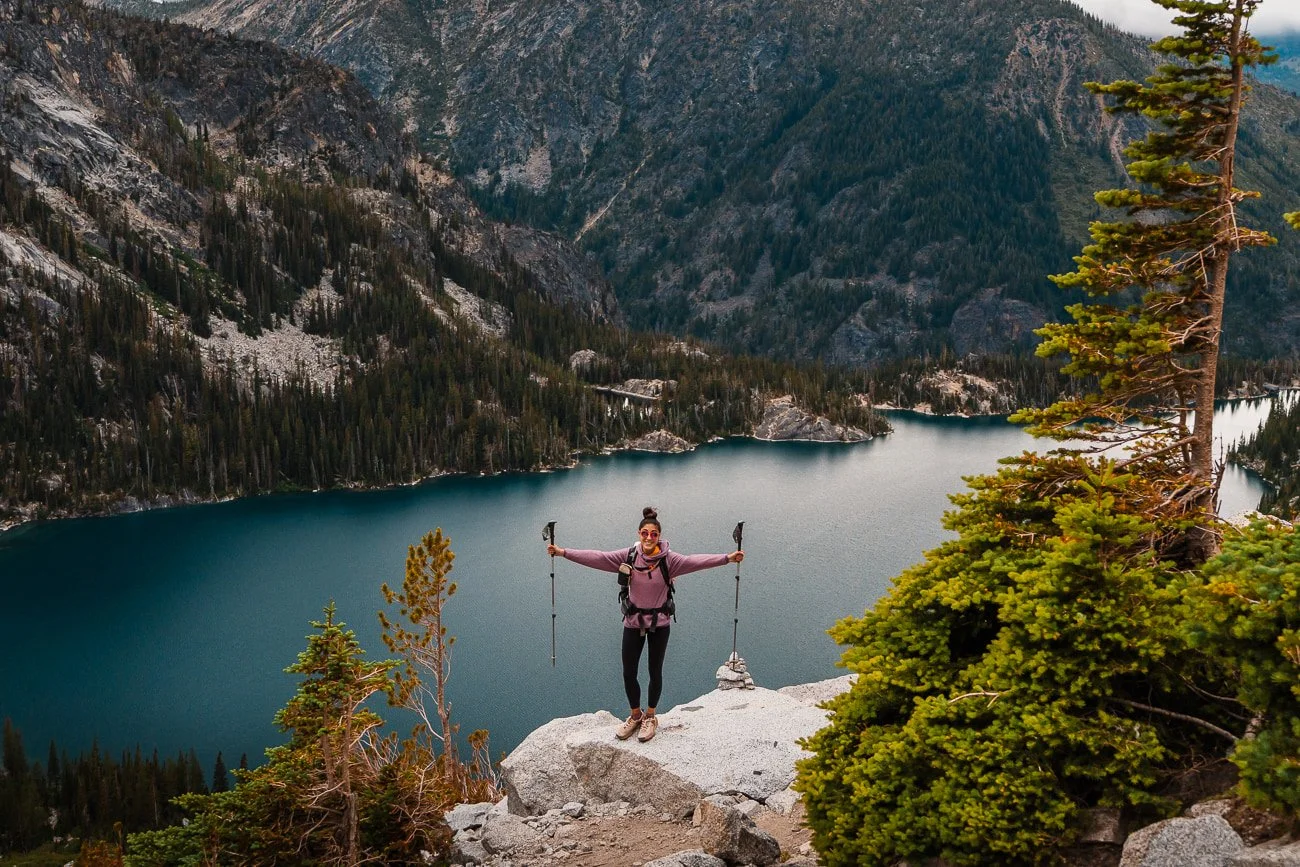 a hiker standing on a rock above a blue lake with arms outstretched