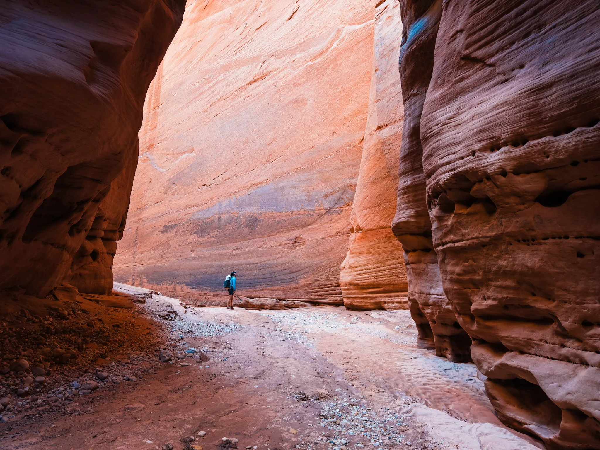 a tiny backpacker, standing in an open section of canyon, looking up at tall and wide sandstone canyon walls
