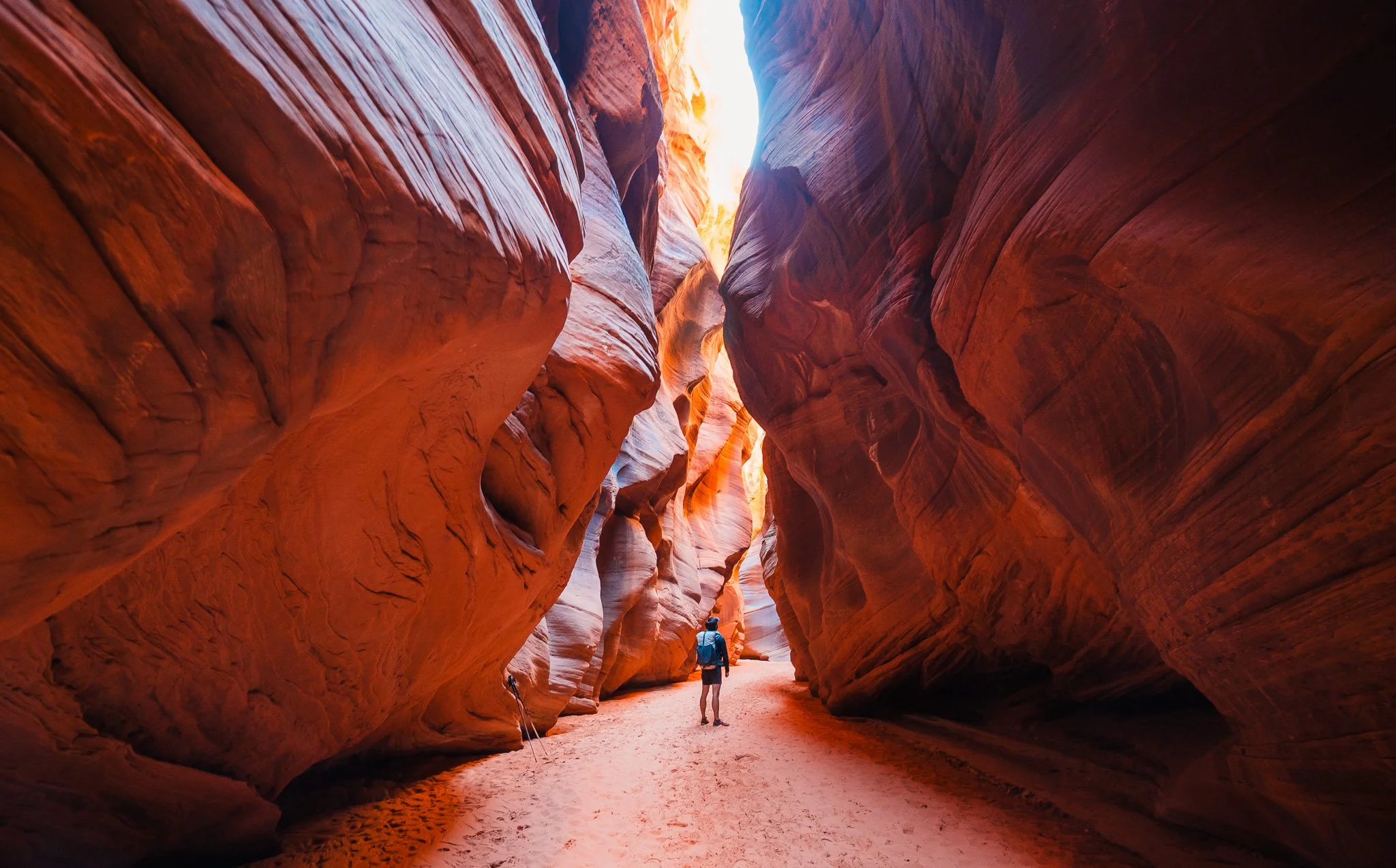 Looking into a slot canyon with high sandstone walls and a sandy trail, with a hiker looking up to the sun