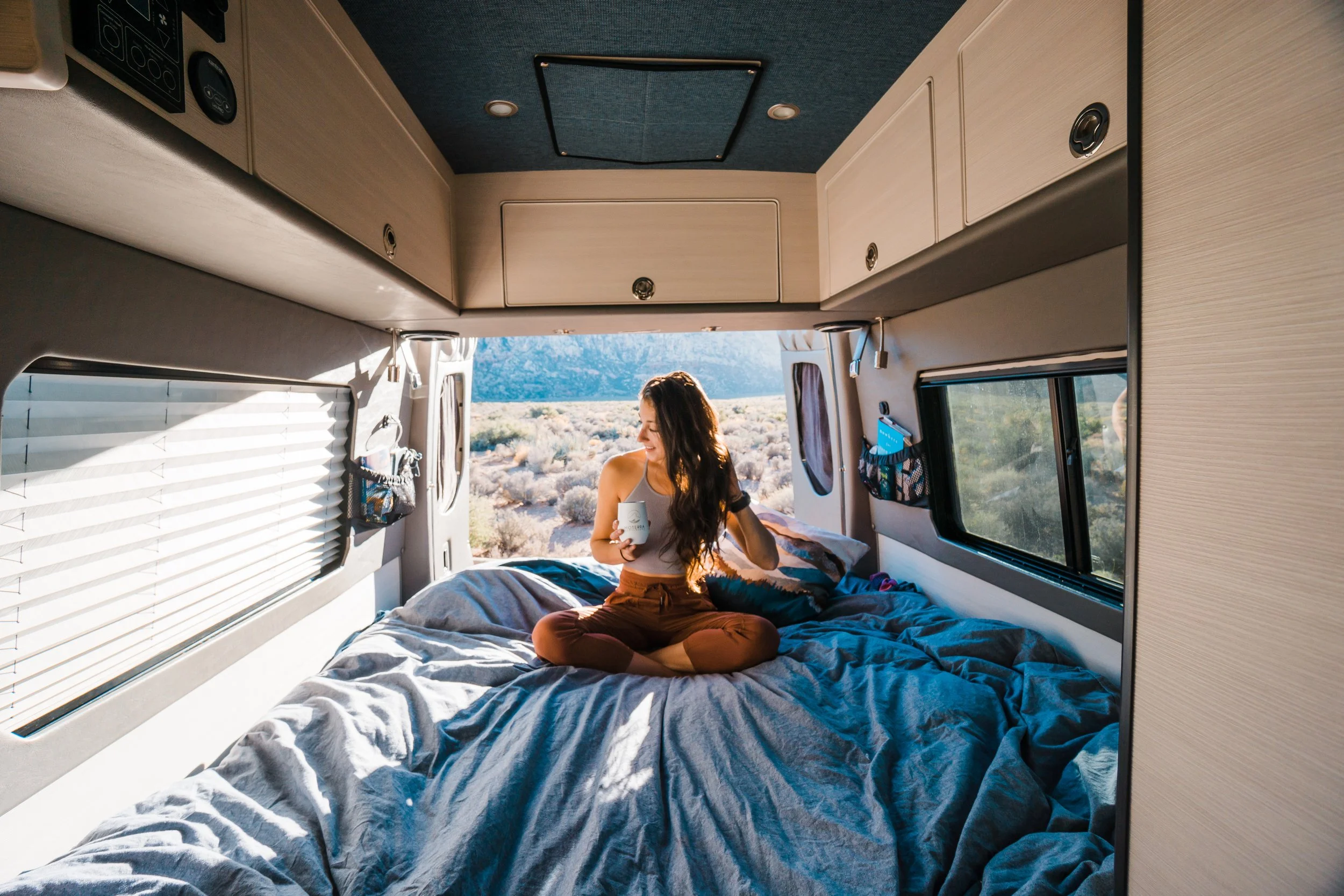 A woman sitting cross-legged on a bed inside a camper van, holding a mug, with desert landscape and mountains visible through the open rear door.