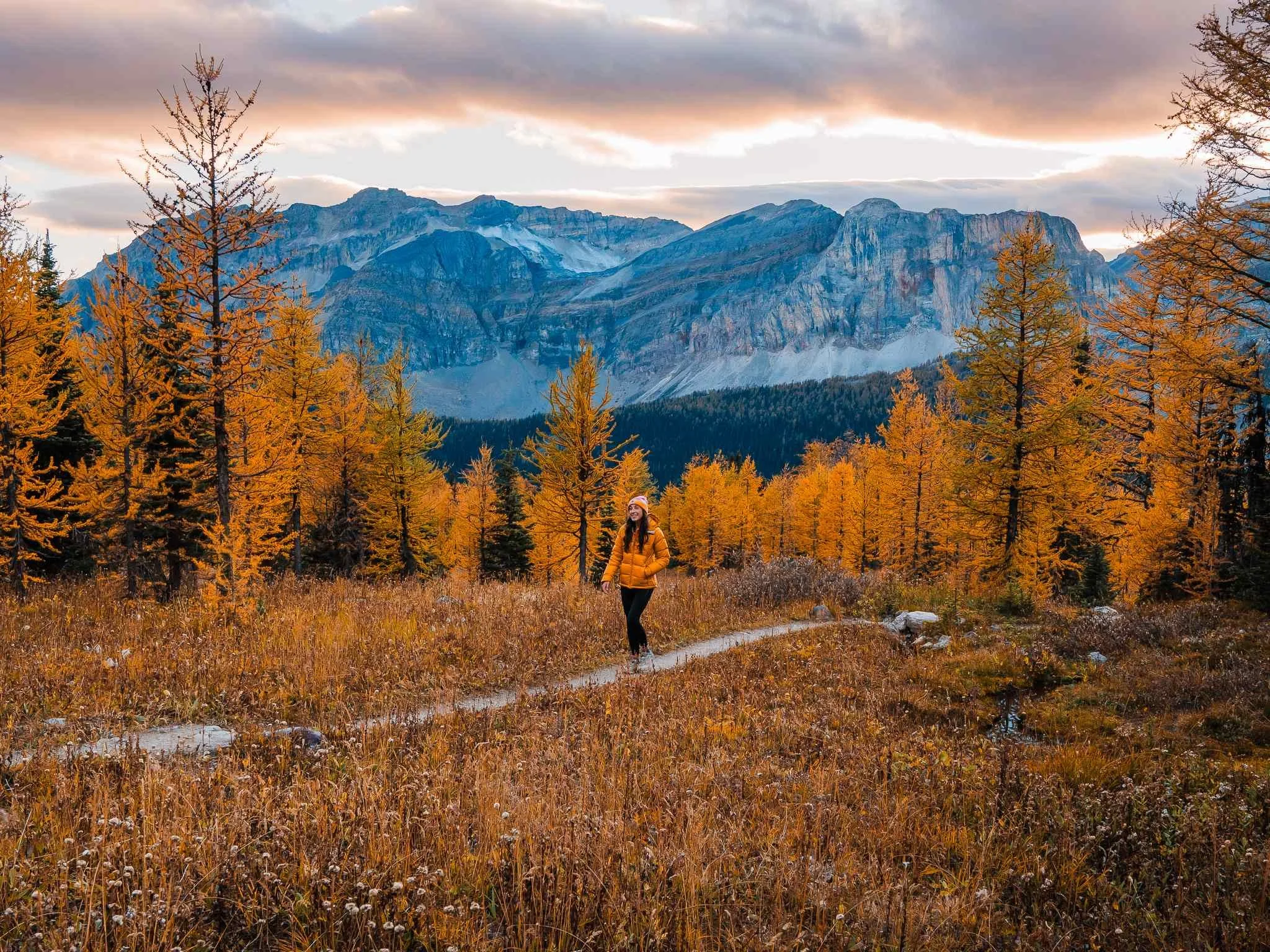 A woman hiking on a trail through an autumn landscape with orange trees, tall mountain range in the background, and a cloudy sky.