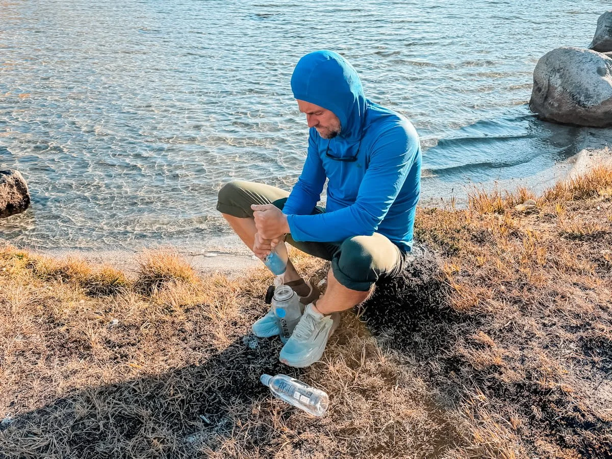 a man in a blue sun hoodie, sitting by a lakeshore in the enchantments, filtering water