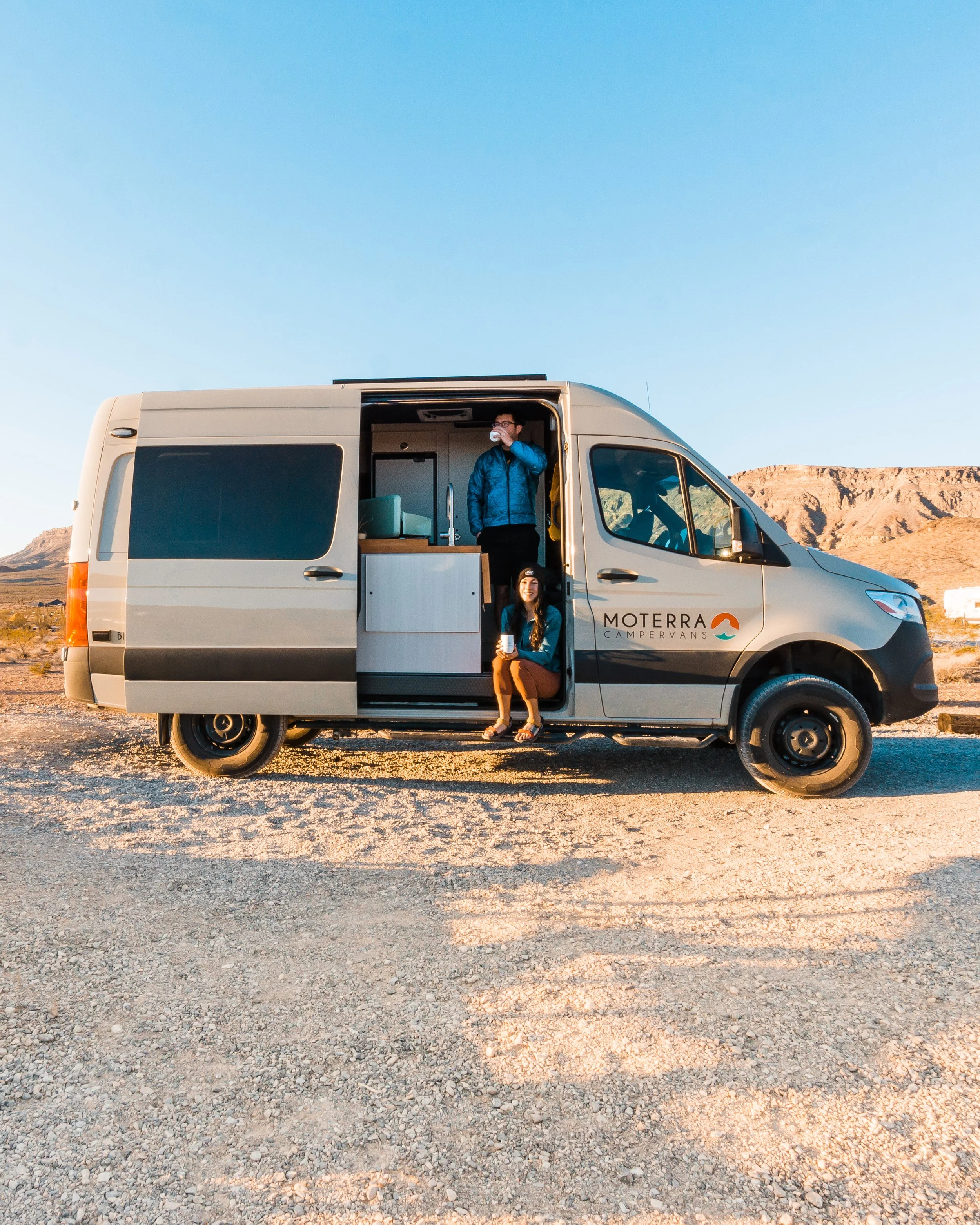 Two people inside and outside a campervan parked on a desert terrain with mountains in the background, during sunset.