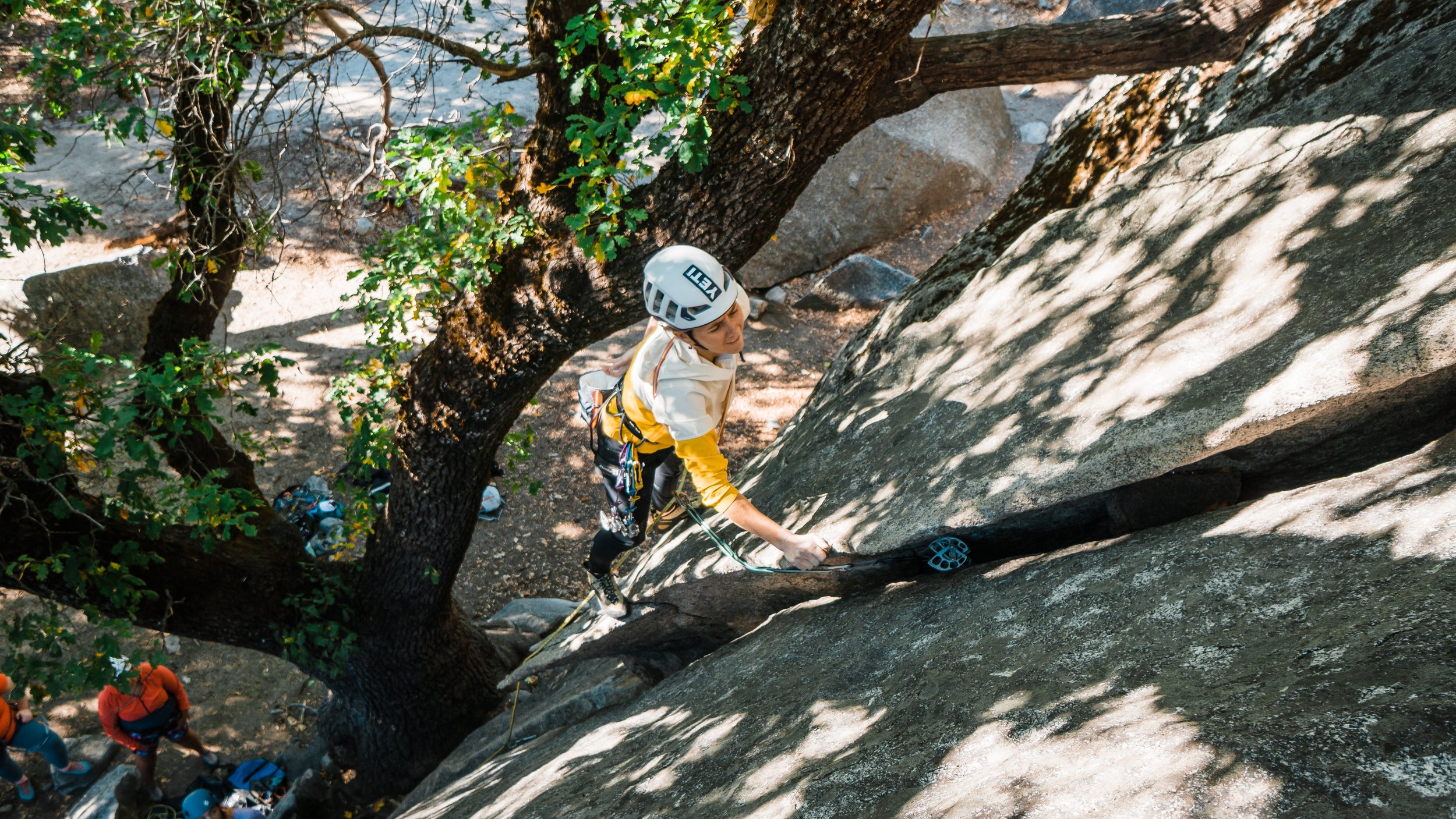 A woman rock climbing outdoors in a helmet and climbing gear, ascending a large granite rock face with trees and other climbers at the base.
