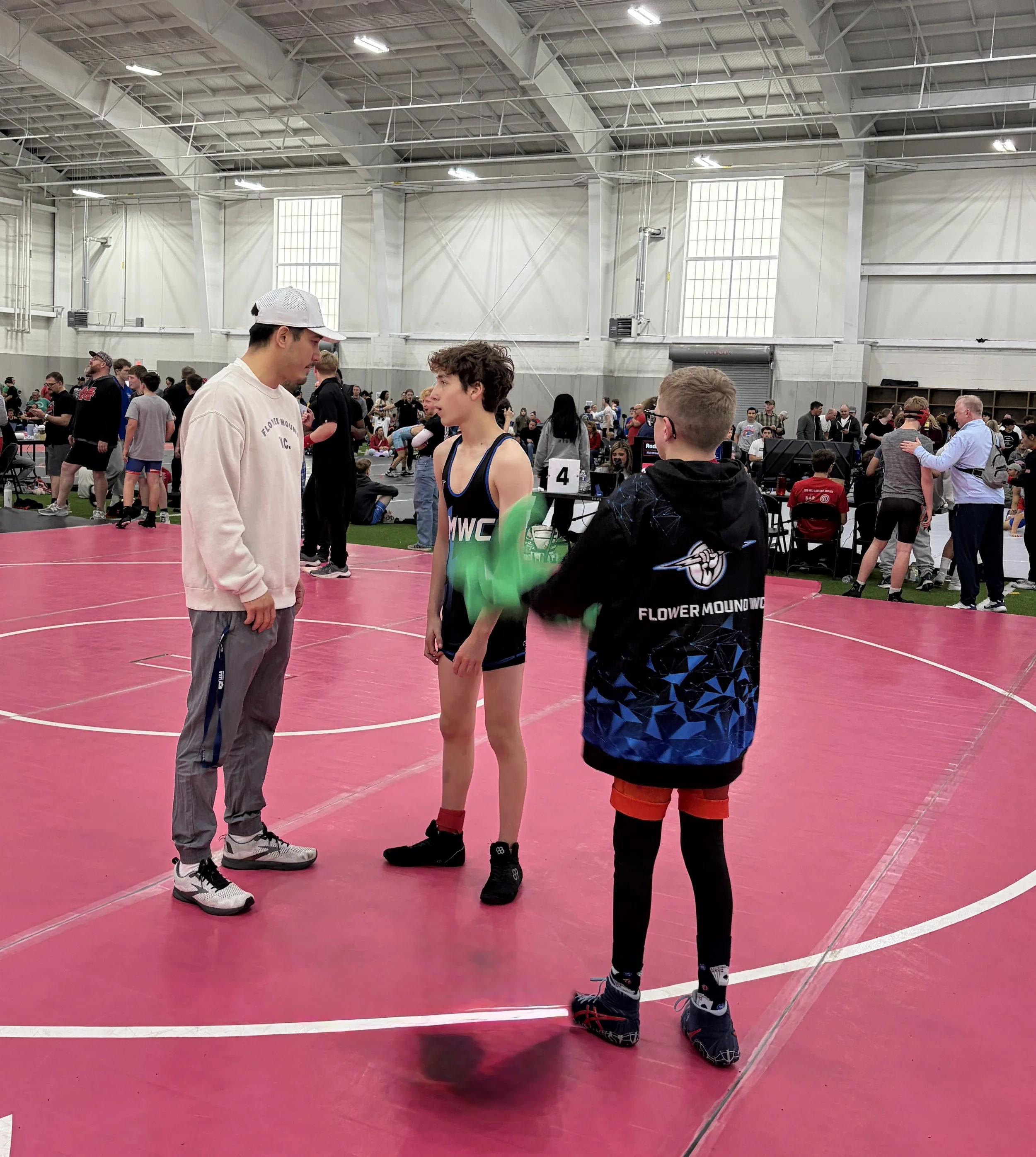 Group of children and adults sitting in a circle on a wrestling mat in a gym, watching two people demonstrate wrestling moves.
