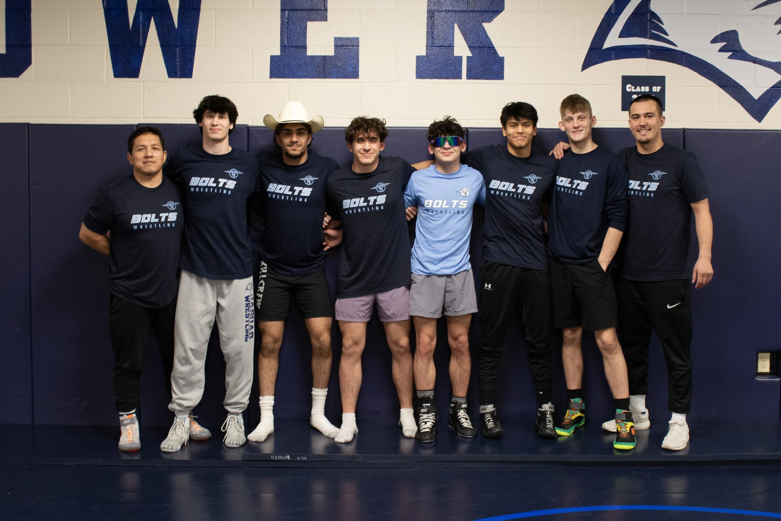 Group of eight young men standing together in a gymnasium, wearing matching 'Bolts Wrestling' shirts, some with shorts and athletic shoes, one with a cowboy hat, smiling with arms around each other.