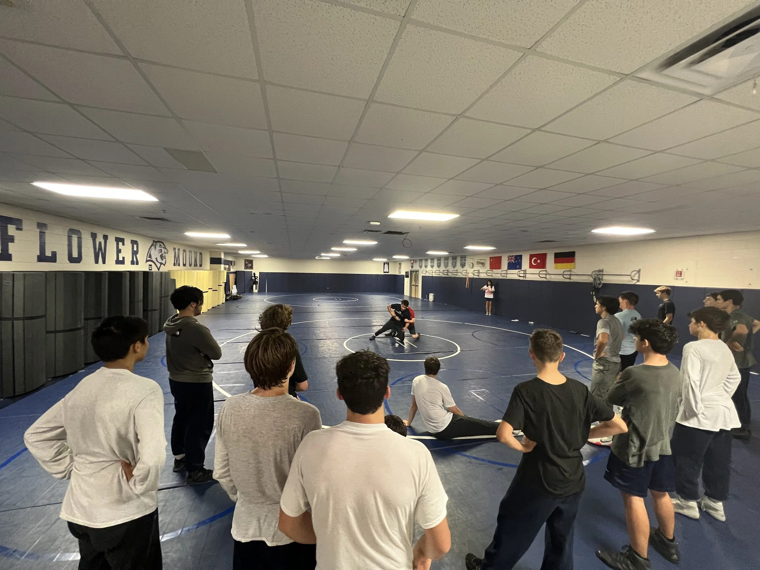 Group of young people observing a wrestling demonstration in a gymnasium with wrestling mats, flags, and banners on the wall.