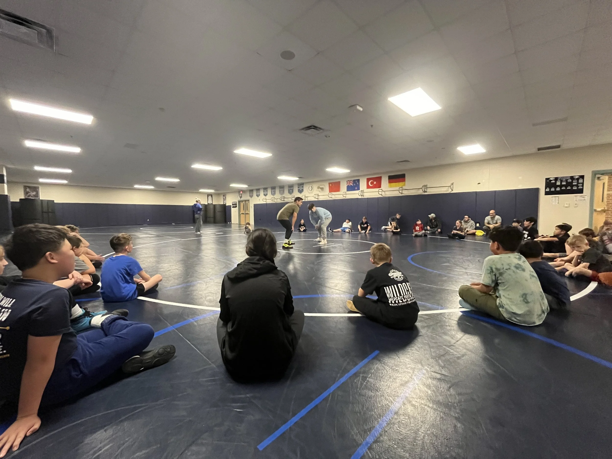Group of children and adults sitting in a circle on a wrestling mat in a gym, watching two people demonstrate wrestling moves.
