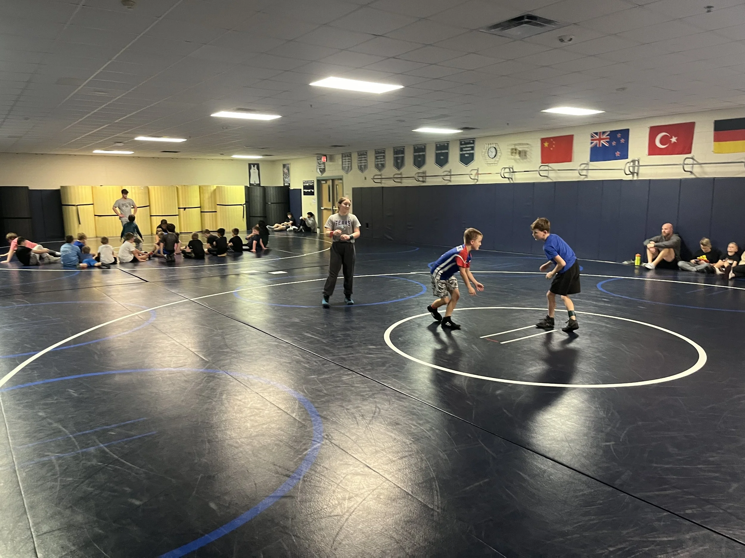 Children practicing wrestling on a black mat in a gymnasium with flags from various countries hanging on the wall.