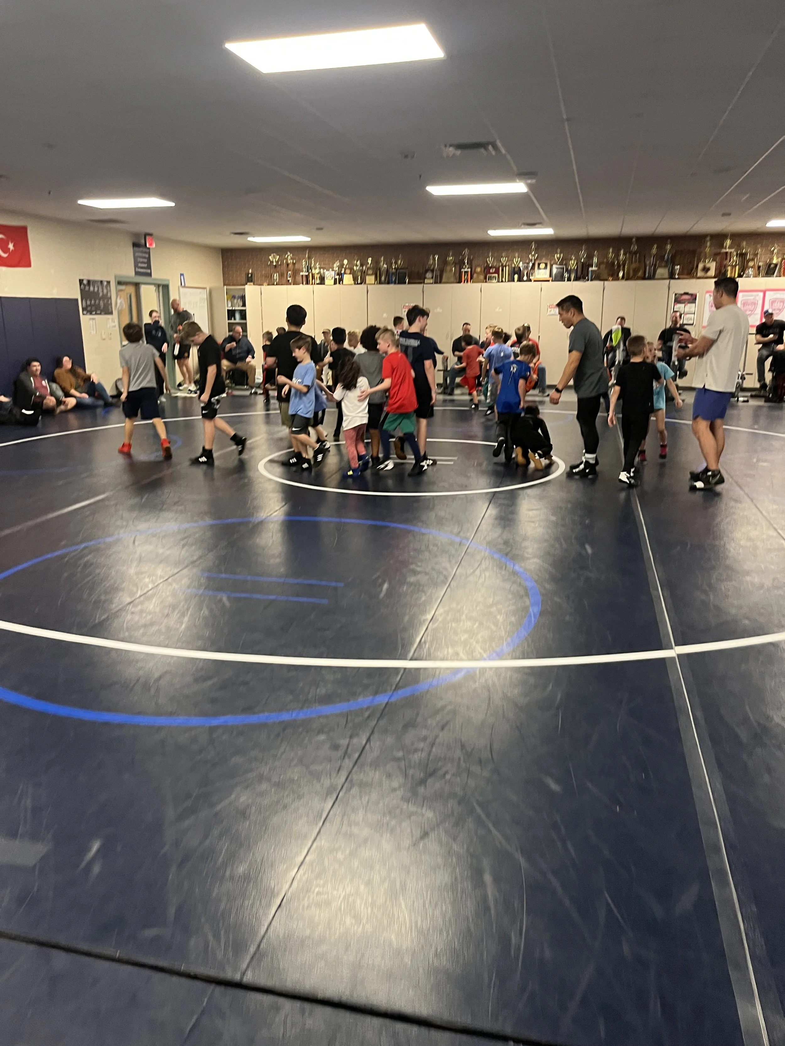 A group of five people, including four young wrestlers wearing medals, poses together at a wrestling event in front of a backdrop with USA Wrestling and United World Wrestling logos. They are smiling, and two adults are kneeling or standing beside the children.