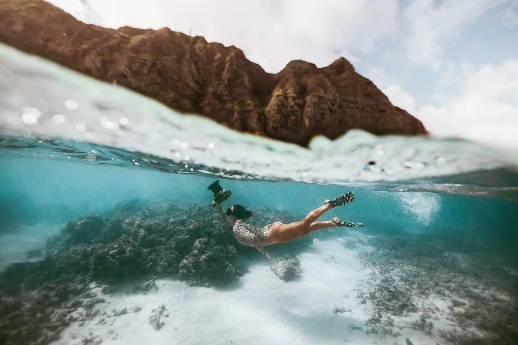 Carol Oliva snorkeling underwater near rocks with a rugged cliff in the background, captured during daytime in the ocean at Oahu Hawaii