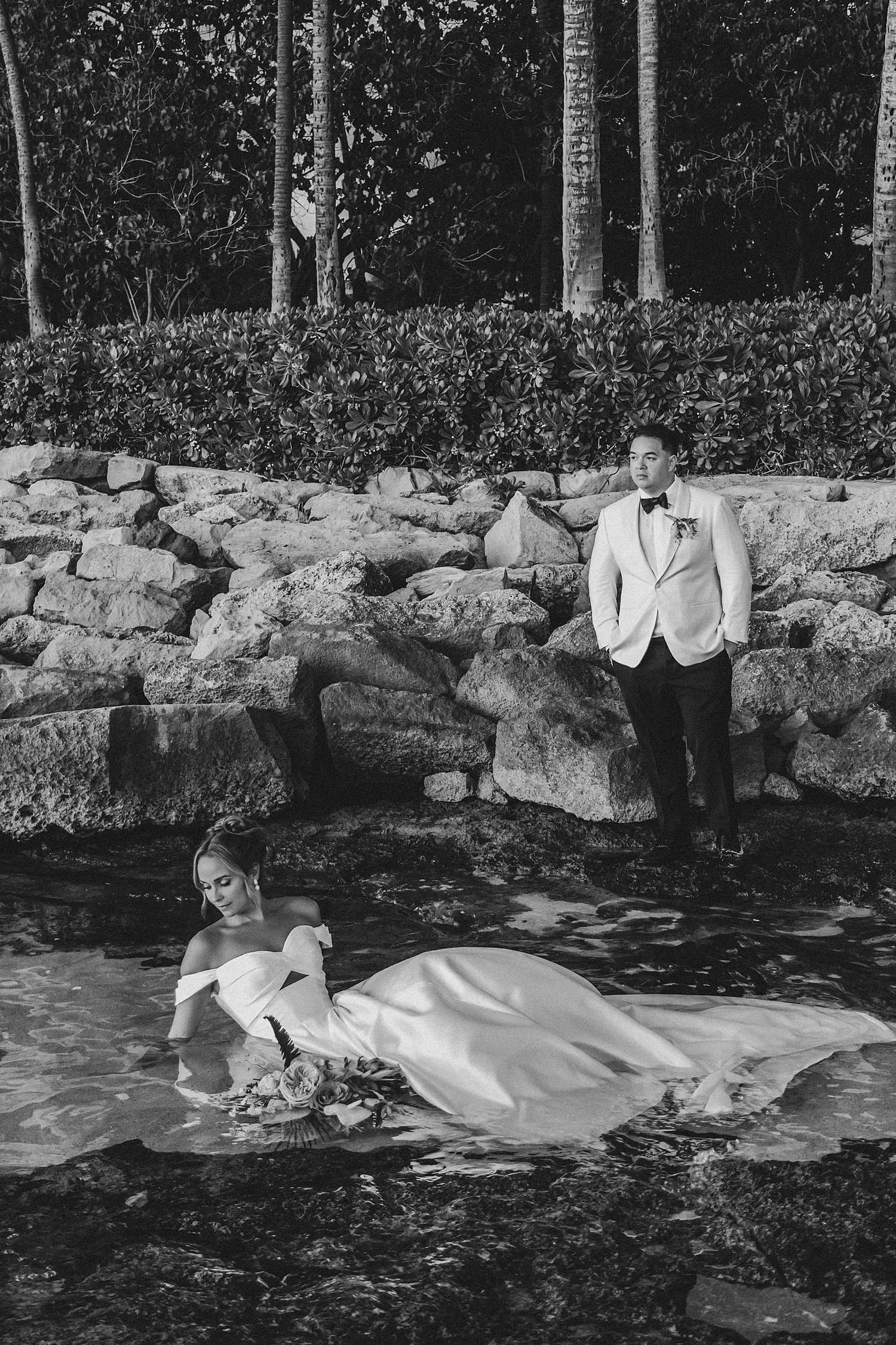     Artistic black-and-white wedding portrait with bride in water and groom standing on volcanic rocks