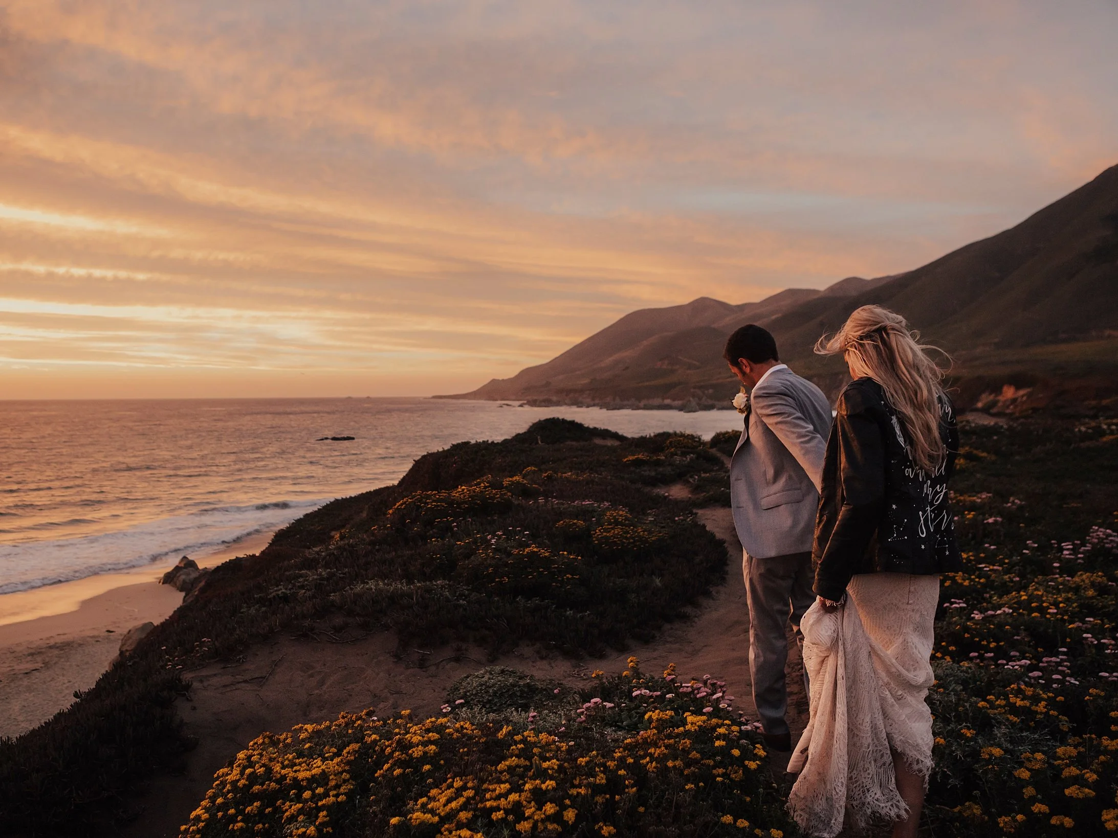 A couple walking along a coastal cliff at sunset, with mountains in the background and the ocean below, as the sky is painted in warm pastel colors.