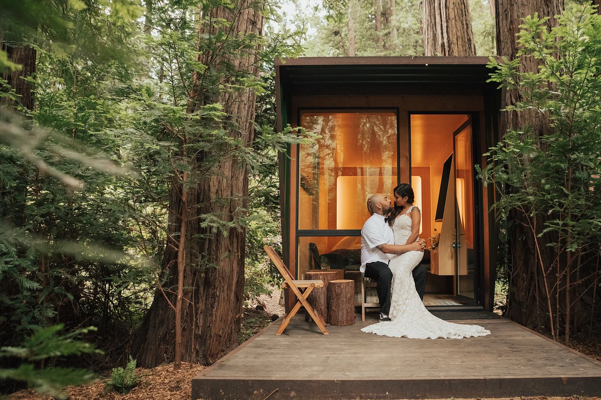 A couple sits inside a small modern cabin surrounded by tall trees, with the man holding the woman's face as they look into each other's eyes, during a wedding or romantic moment during the day.
