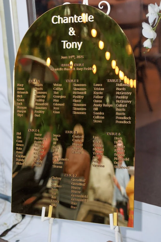 Gold mirror wedding seating chart displayed at In The Grove in Bundaberg, showing guest table assignments with warm fairy light reflections and elegant styling.”