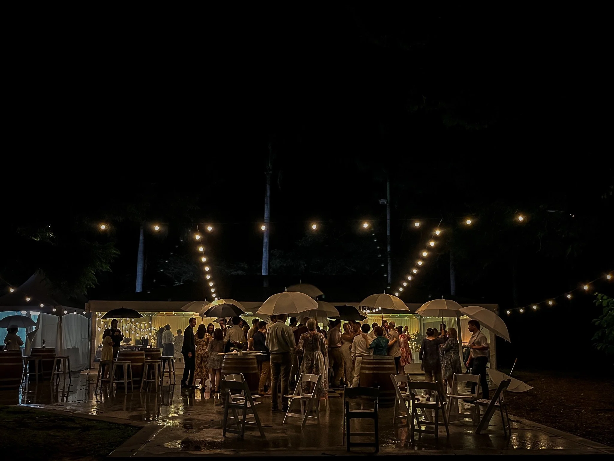 Wedding guests gathered beneath umbrellas on a rain-wet patio at In The Grove, with festoon lights glowing overhead.