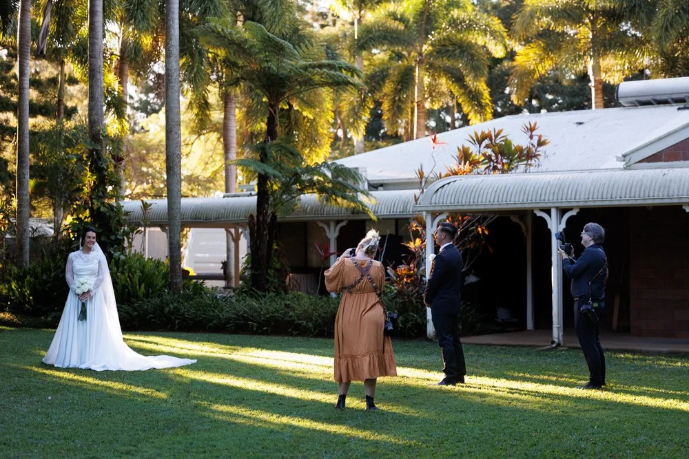“Bride posing in the garden at In The Grove wedding venue in Bundaberg while photographers capture portraits among the palm trees and soft afternoon light.”