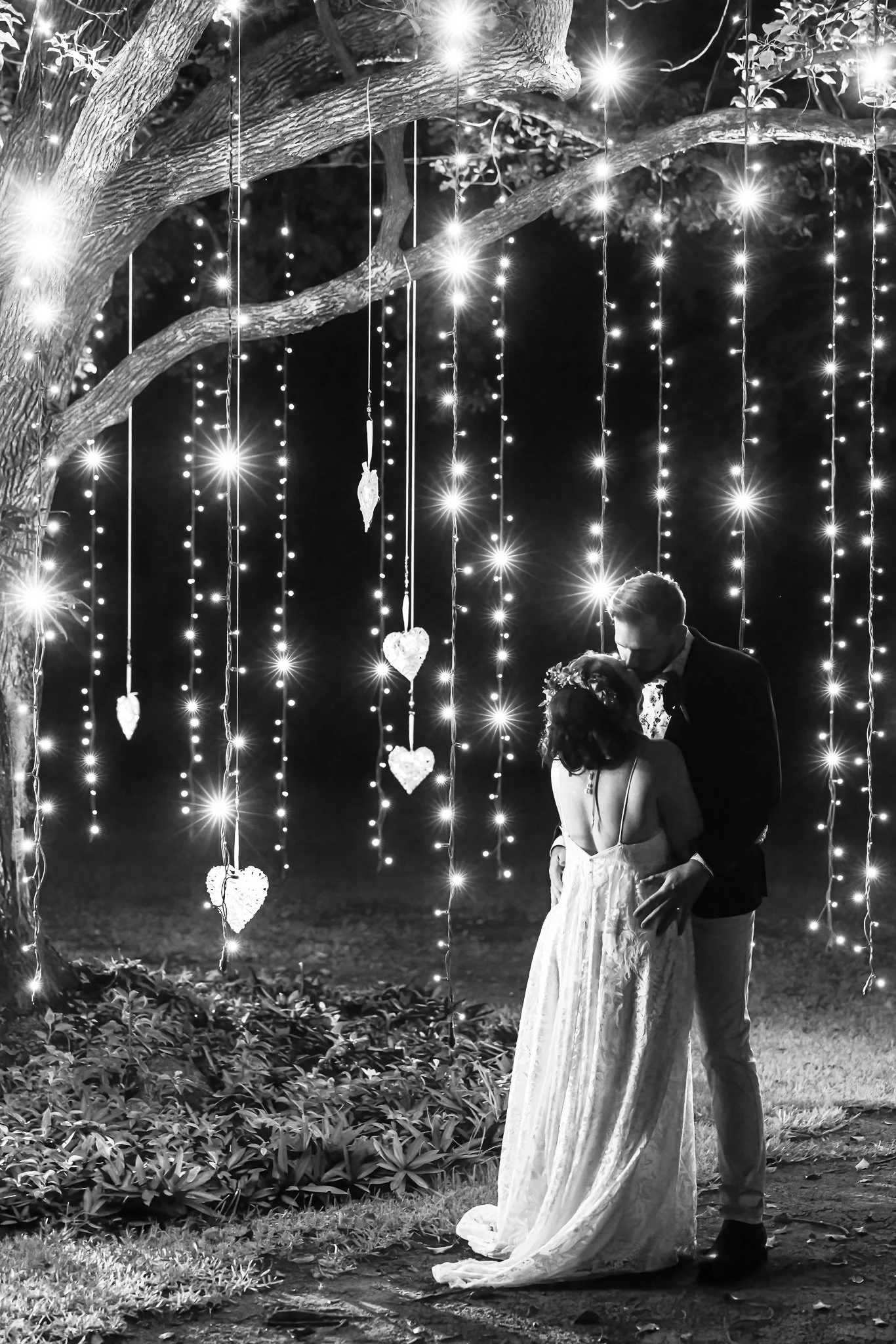 Bride and groom sharing a romantic moment beneath a tree draped in fairy lights at night.