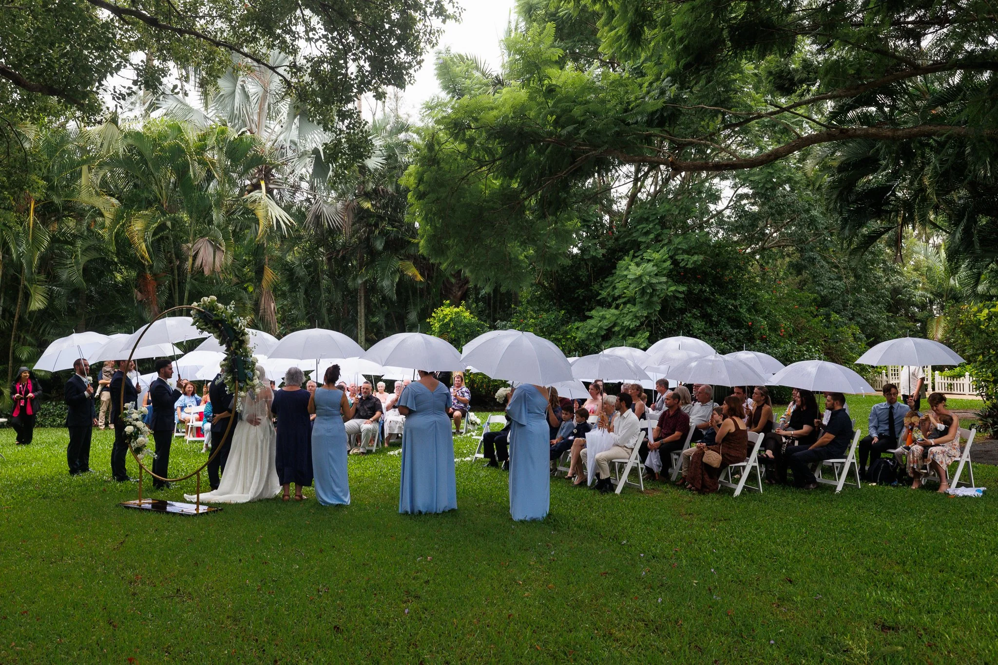 Wedding couple exchanging vows in the gardens at In The Grove, with guests and the bridal party sheltering beneath white umbrellas.