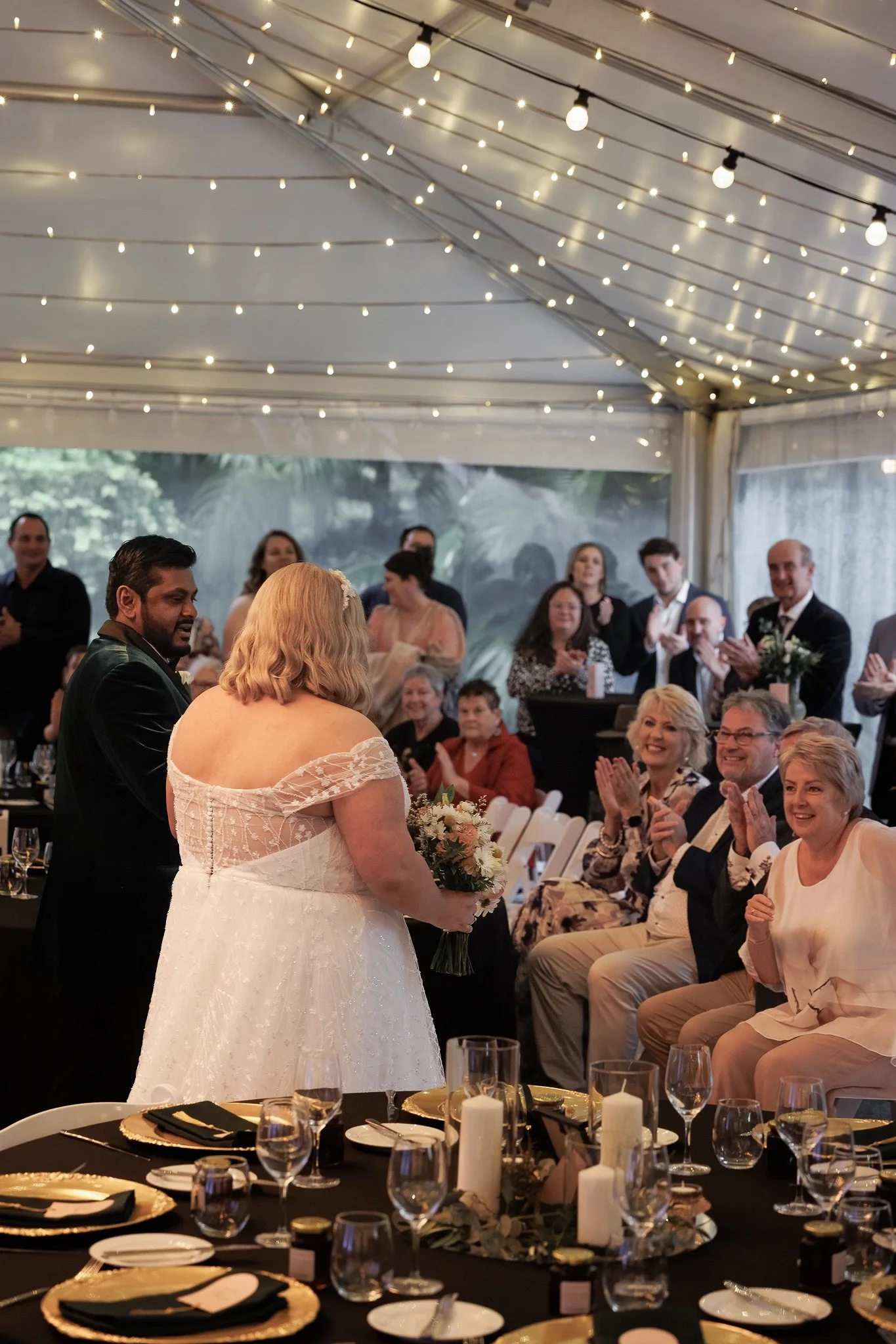 Wedding couple surrounded by smiling guests inside In The Grove’s reception marquee, beneath strings of warm lights.