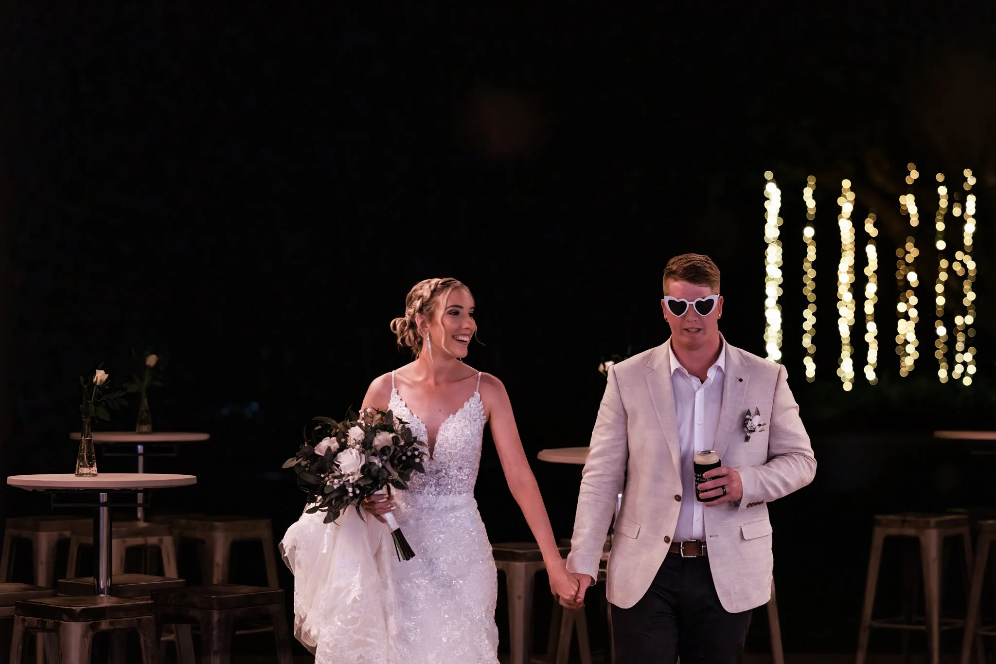 Bride and groom entering their wedding reception at night with playful energy and glowing lights behind them.