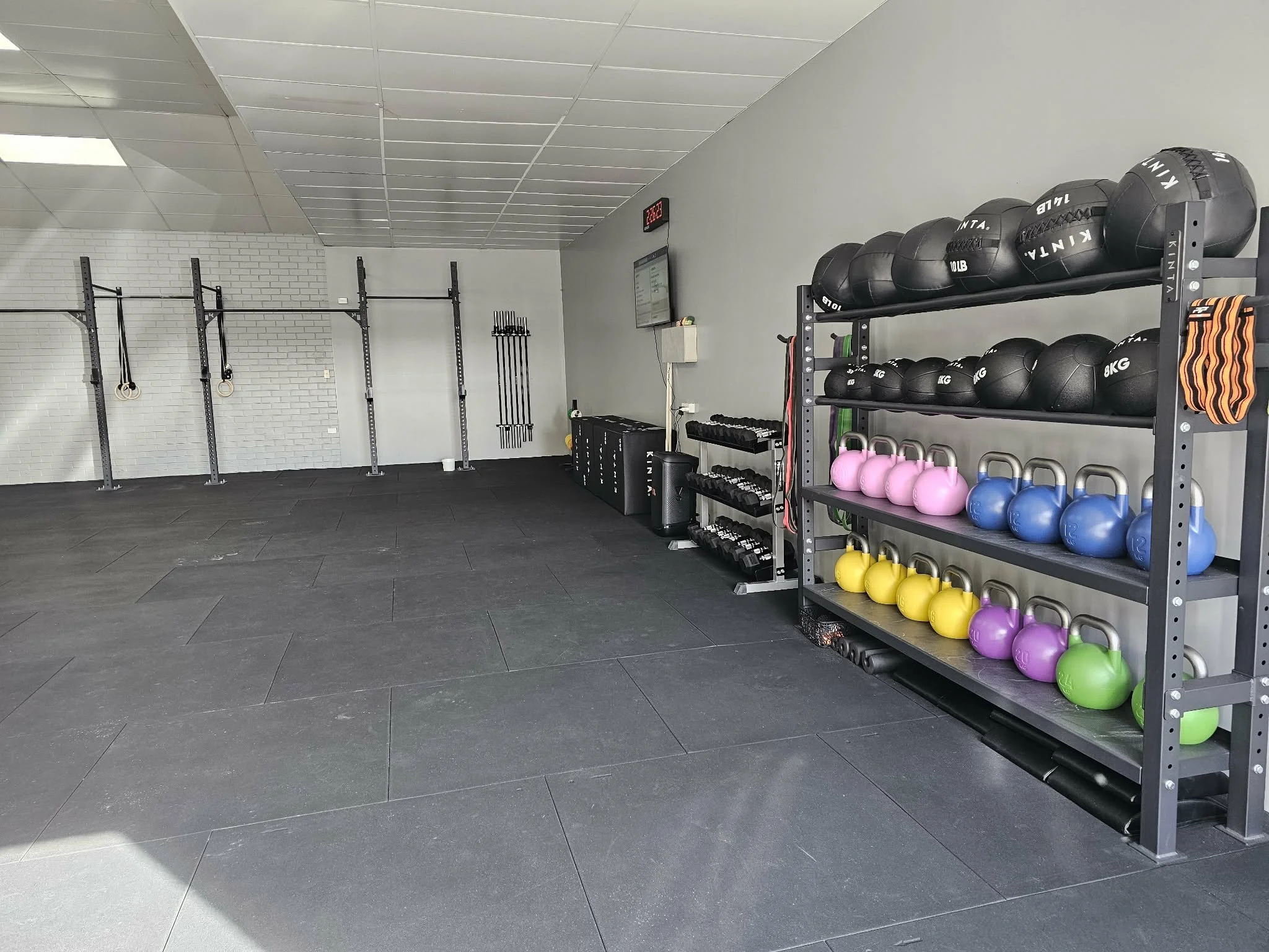 Empty gym with black rubber flooring, white brick wall, and workout equipment including kettlebells in various colors and weights, medicine balls on a shelf, and gymnastic rings hanging from the wall.