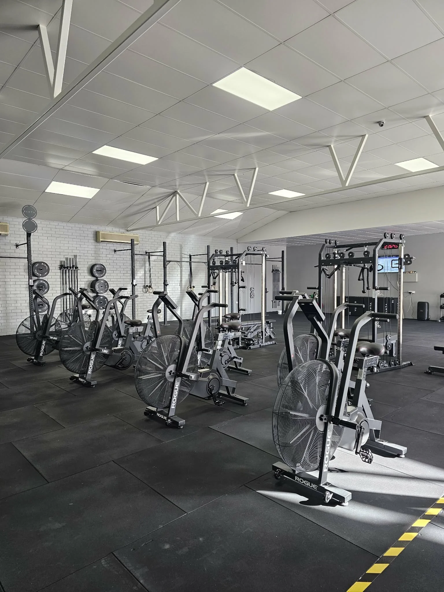 Empty gym with row of ngeellike exercise bikes and weightlifting equipment on black rubber flooring.