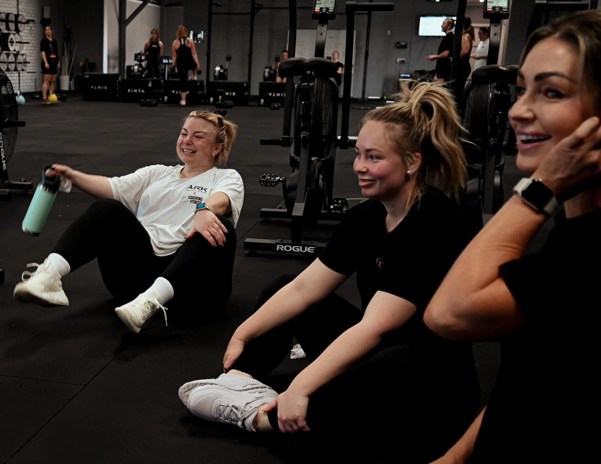Three women sitting on the floor of a gym, smiling and laughing, with exercise equipment and other people working out in the background.