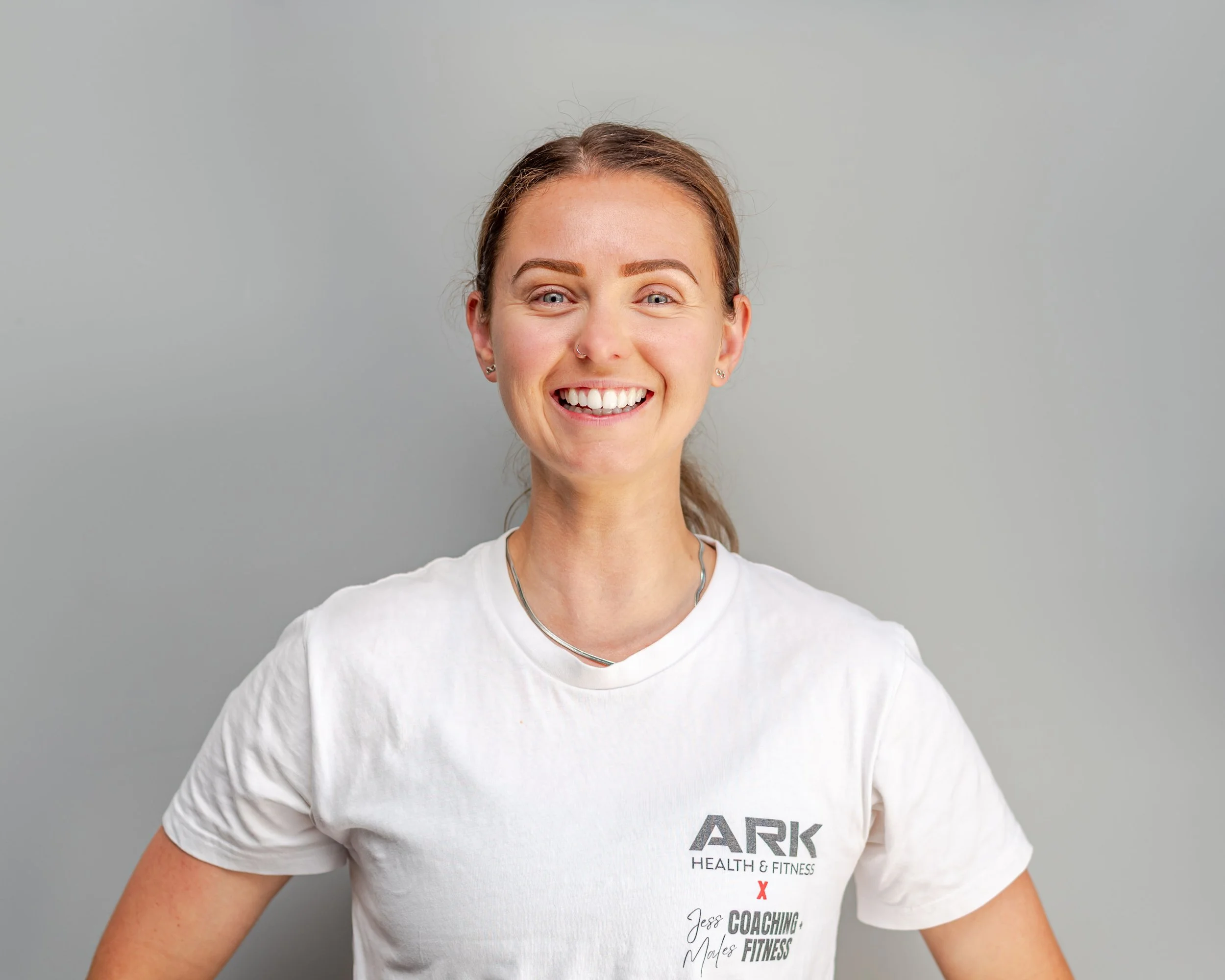 A smiling woman with brown hair tied back, wearing a white t-shirt with 'ARK HEALTH & FITNESS' and 'Jens Coaching, Males Fitness' printed on it, standing against a plain gray background.