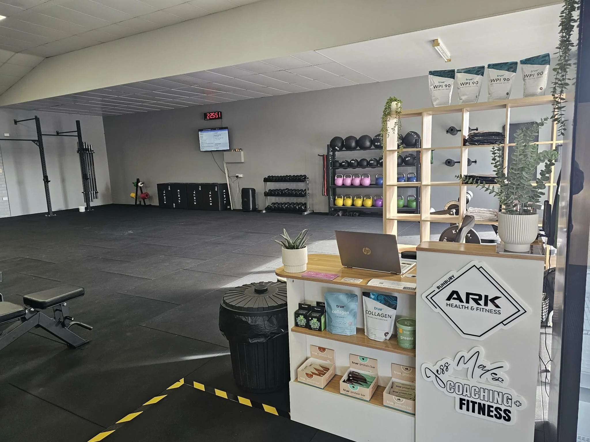 Empty fitness gym with weightlifting equipment, kettlebells, and a reception desk with fitness supplements, plants, and a digital clock and screen on the wall.