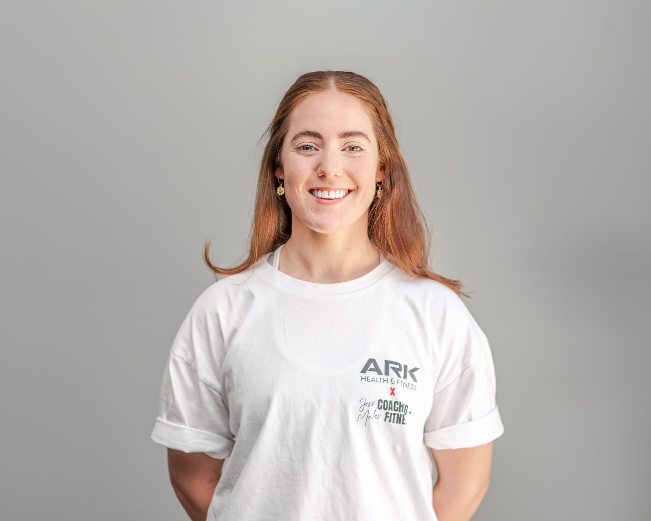 A young woman with long red hair smiling, wearing a white T-shirt with black and red text, against a plain gray background.