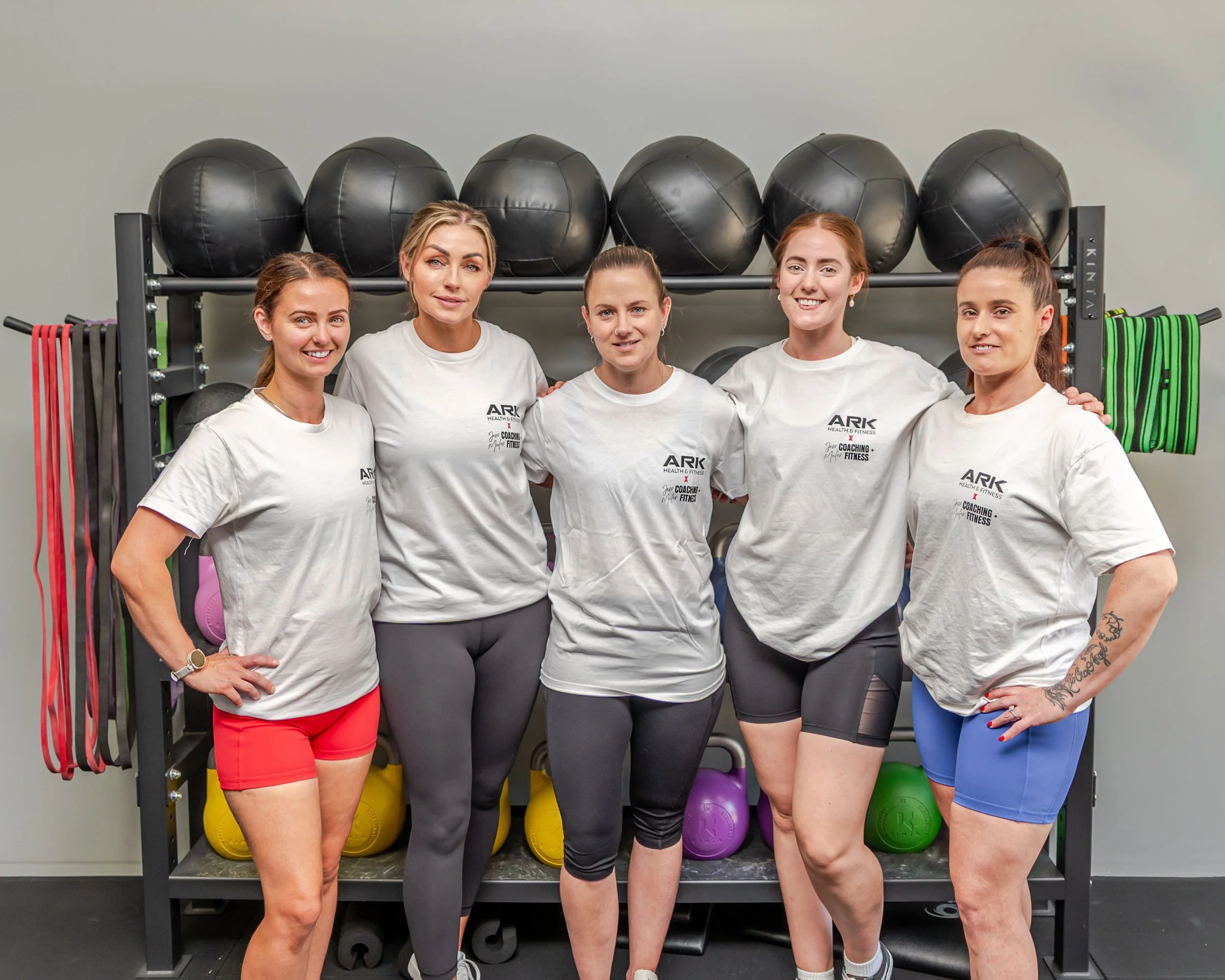 Group of five women in workout clothes standing together in a gym, smiling. They are in front of exercise equipment including medicine balls, kettlebells, and resistance bands.