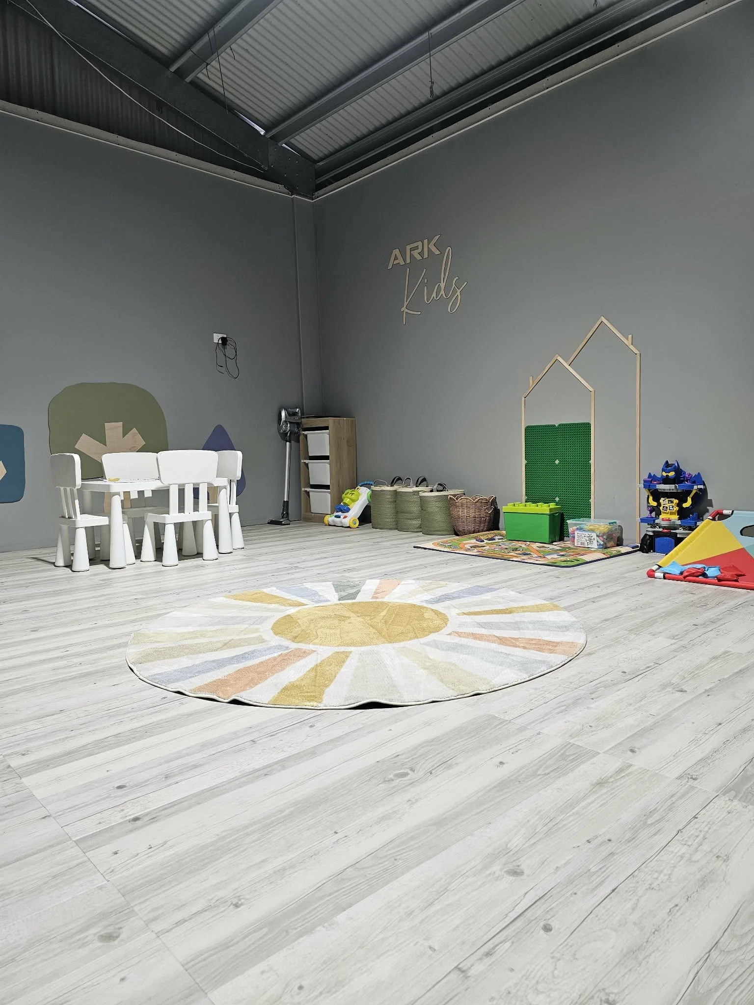 Empty kids' playroom with light wood flooring, a round sunburst patterned rug, small white table with chairs, storage baskets, toys, and wall decor reading 'ARK Kids'.