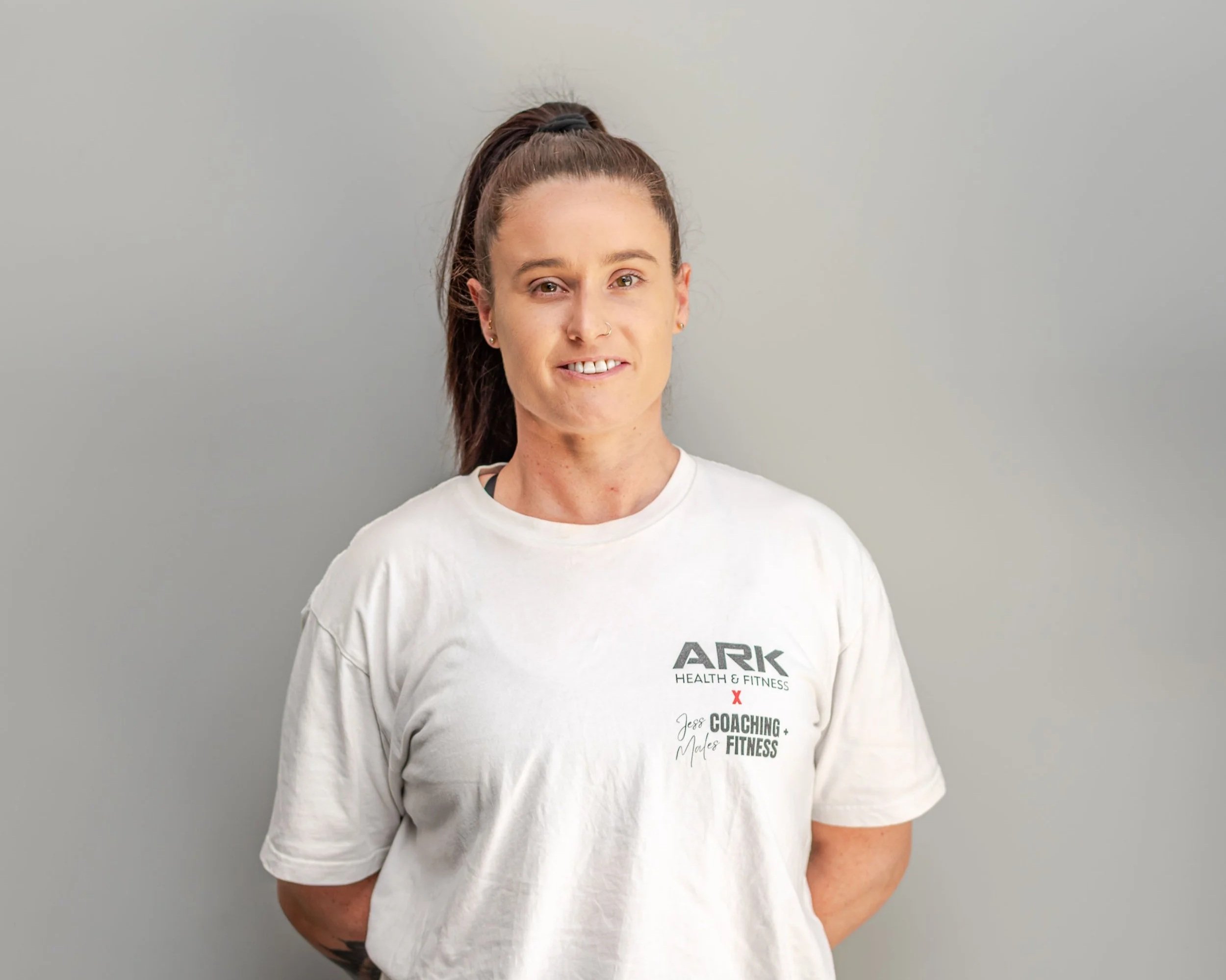 Woman with long dark hair tied in a high ponytail, wearing a white T-shirt with 'ARK HEALTH & FITNESS' text, standing against a plain gray wall.