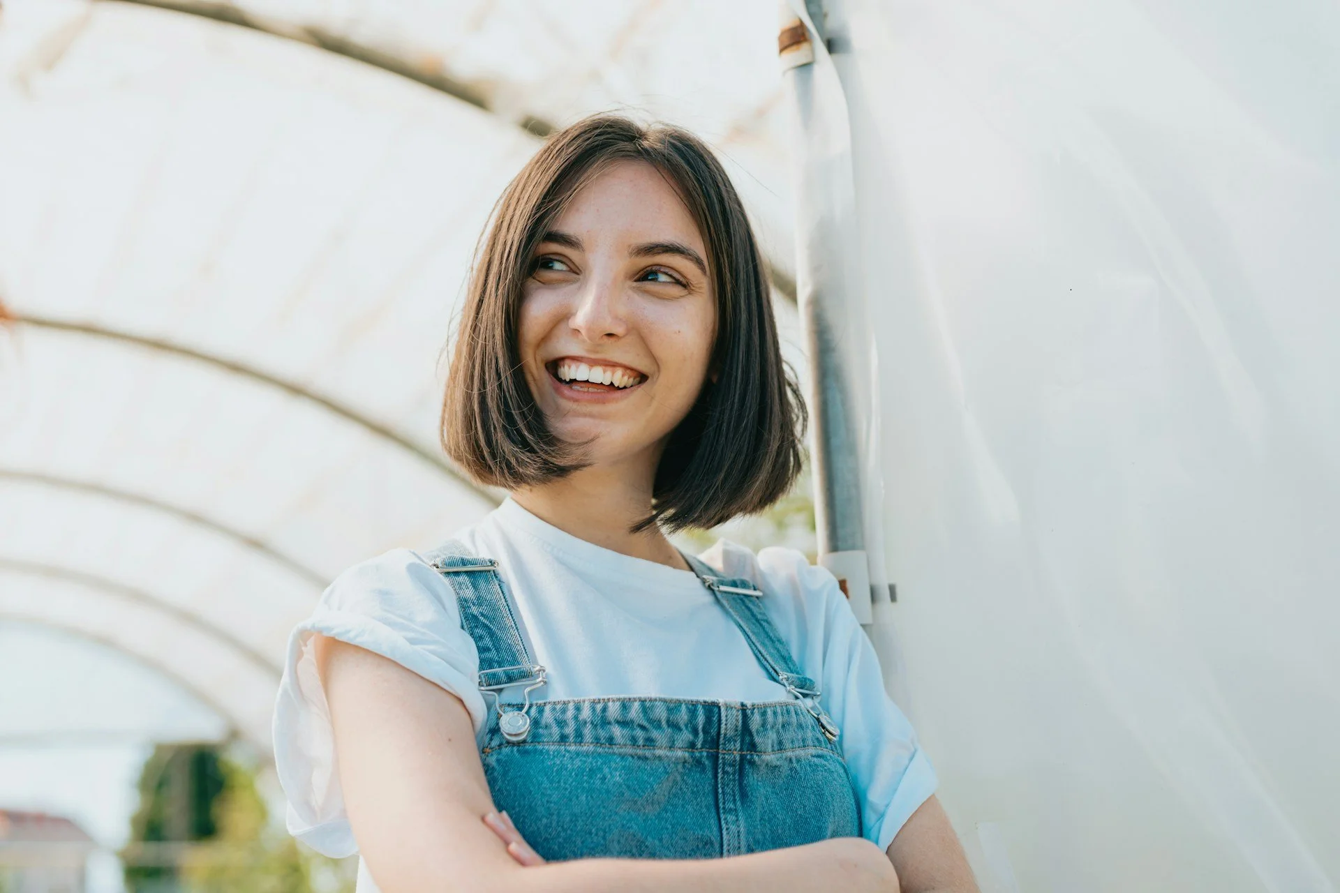 Young woman in overalls has arms crossed and is smiling.