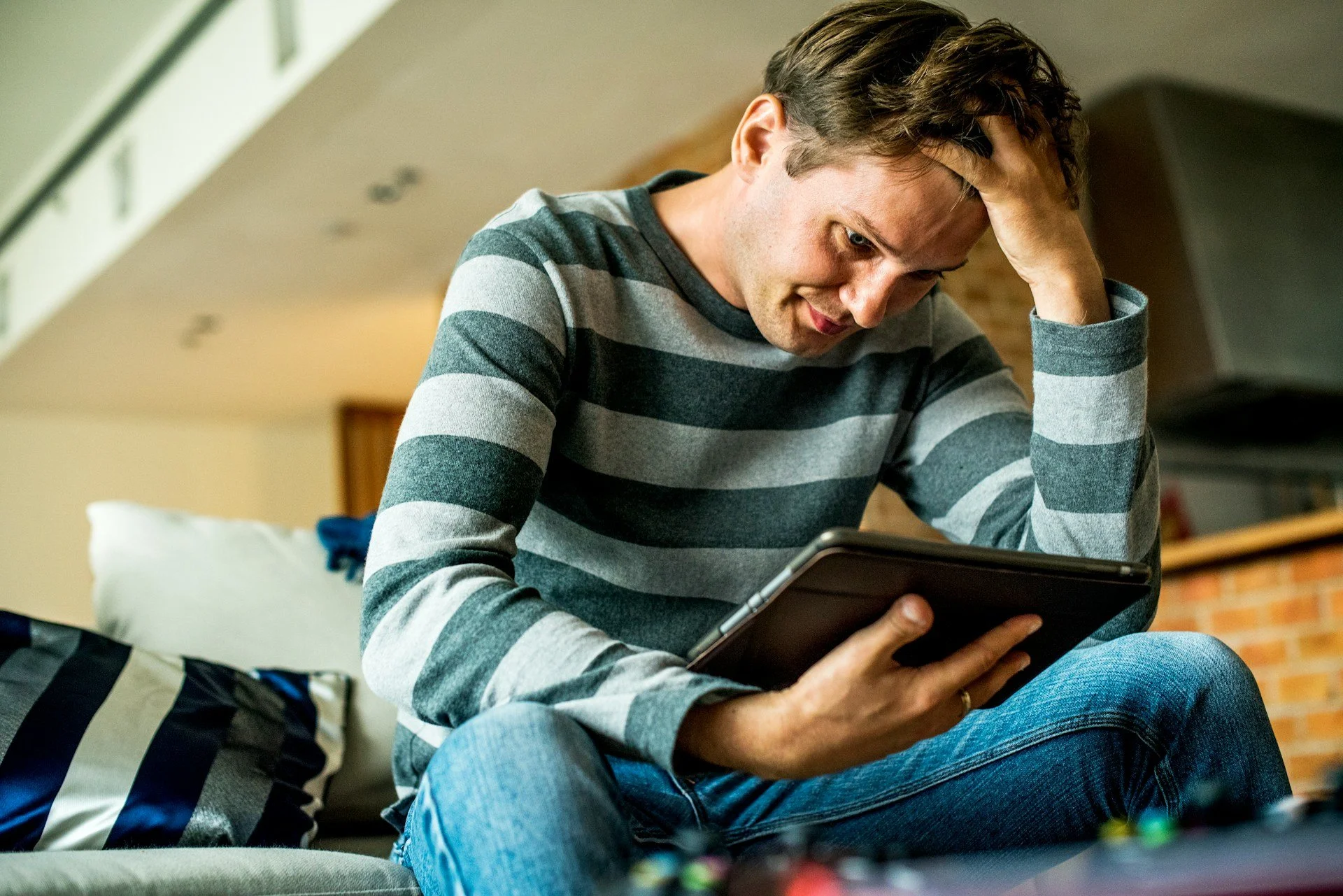 Young man is looking at a tablet, with his hand on his head, appearing stressed.