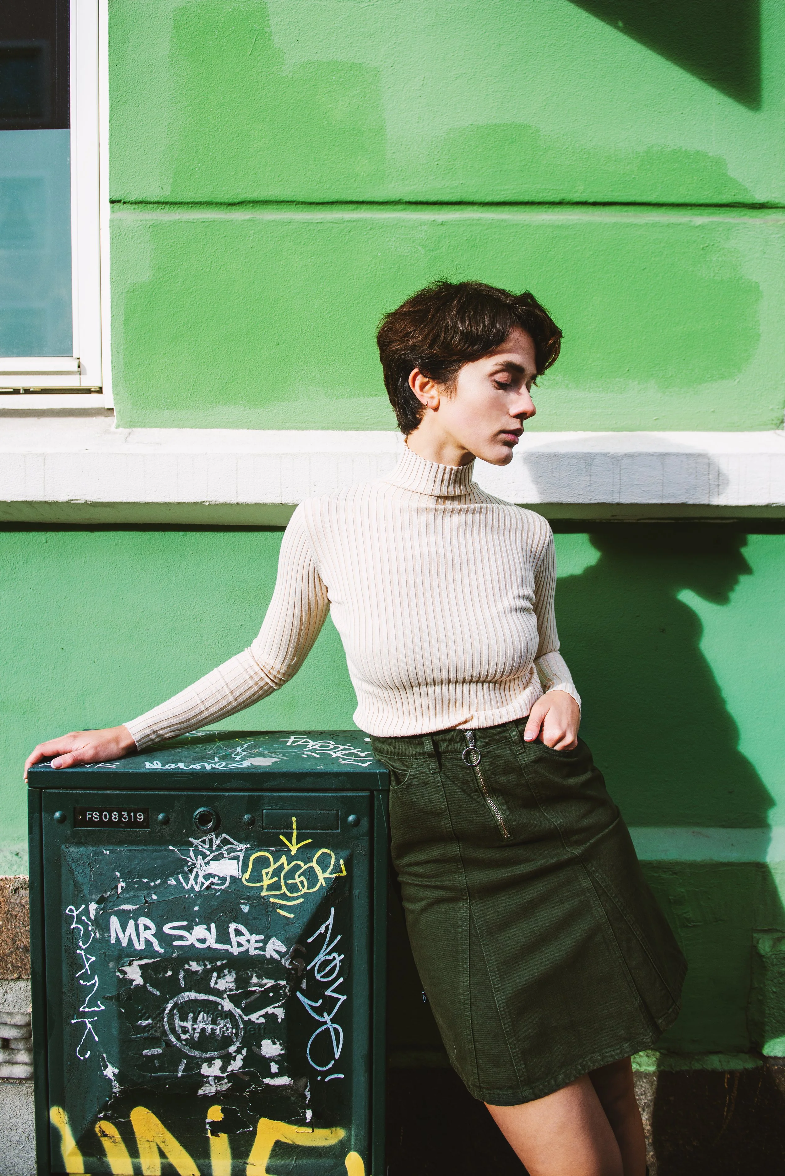 susanne karin moe leaning against a metal box out on the street with a bright green wall behind her