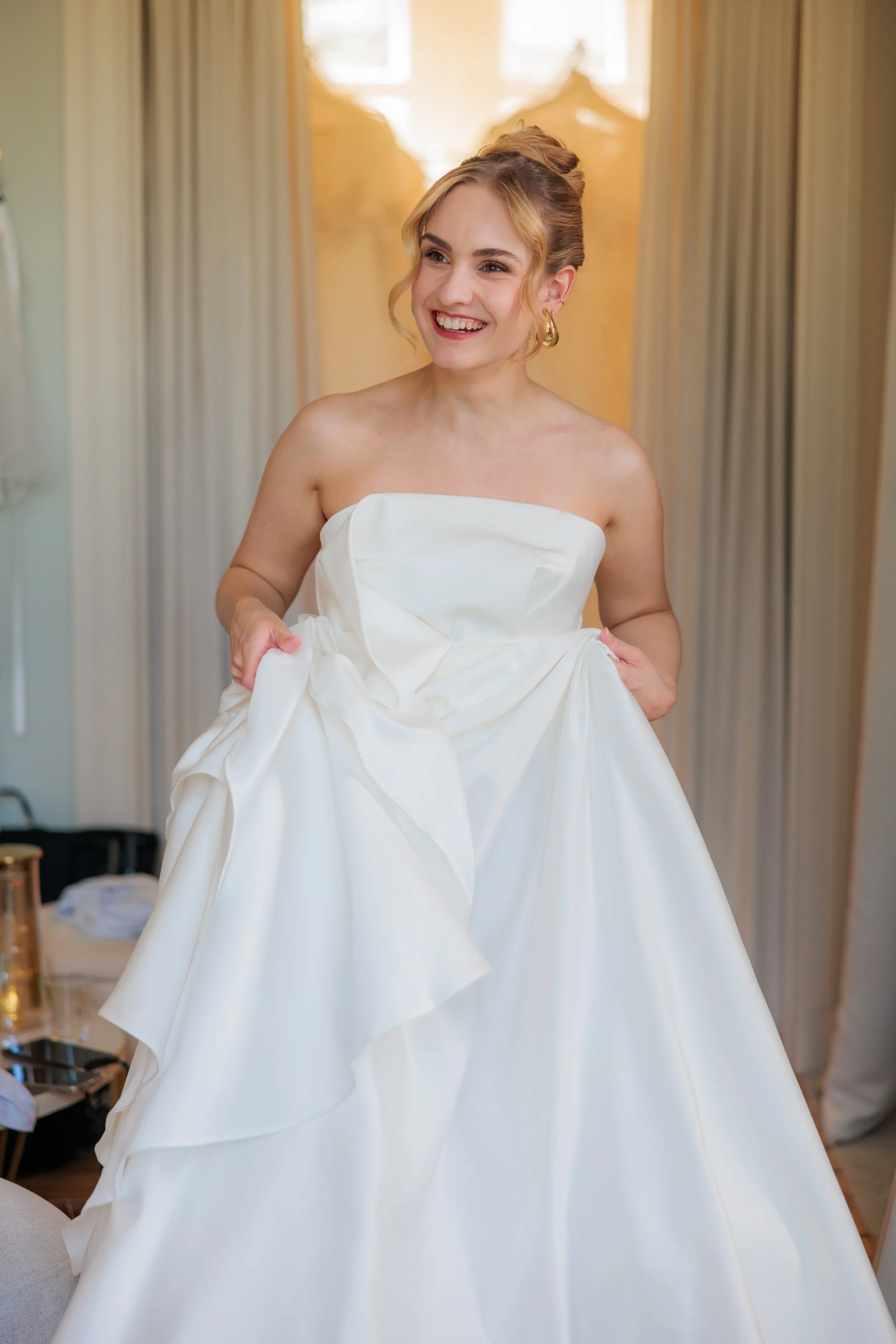 A woman smiling in a strapless white wedding dress, holding the skirt of the dress, standing indoors with sunlight coming through the window in the background.