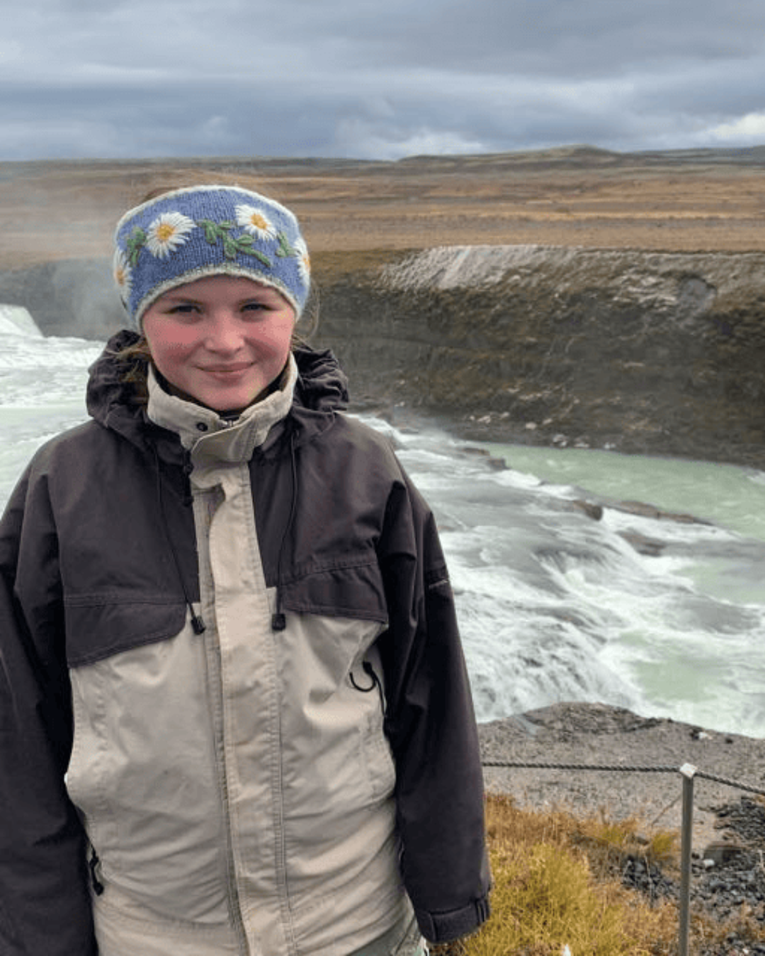 A girl with brown hair and a daisy headband standing in front of a waterfall.