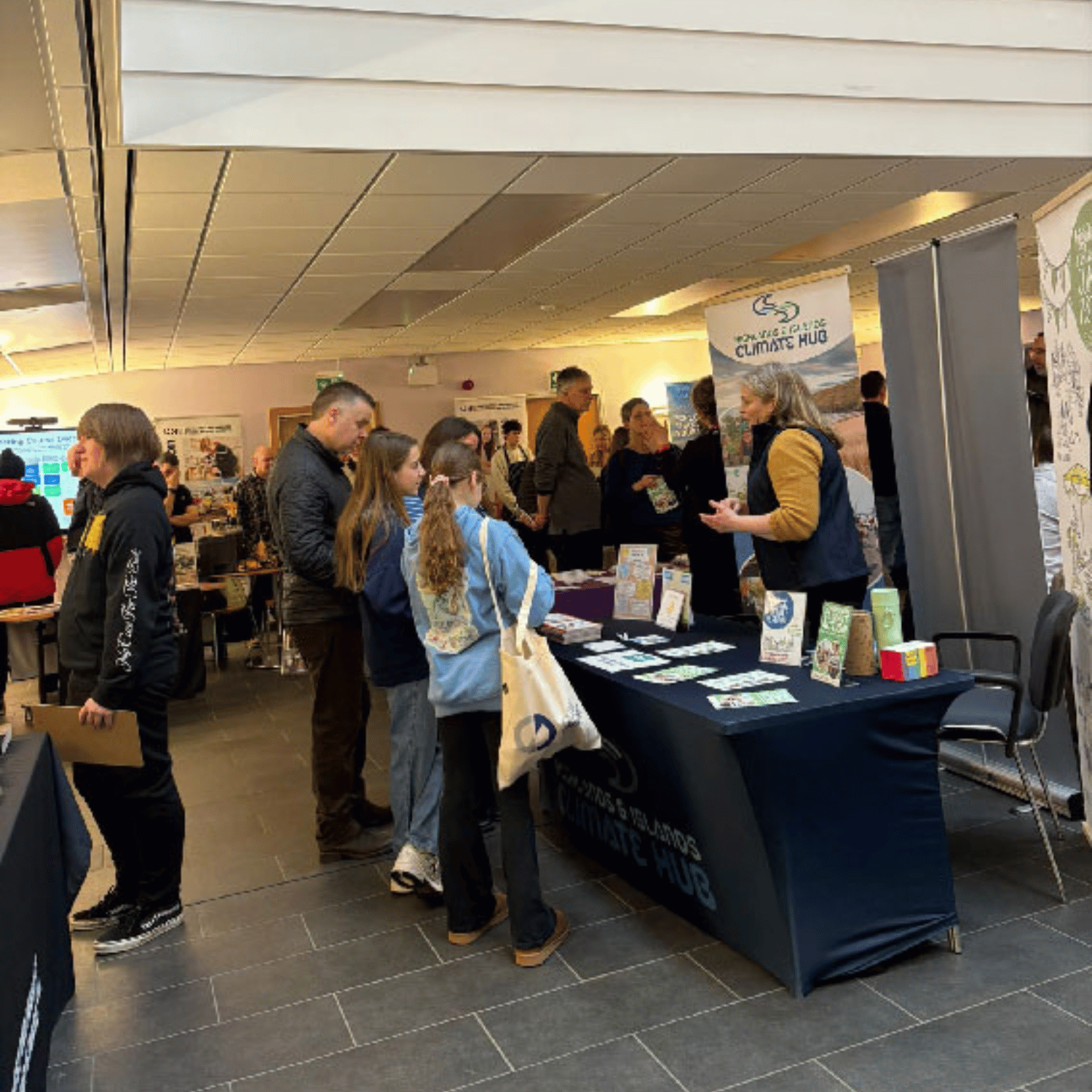 A father and his two daughters talking to the Highlands & Islands Climate Hub at a recruitment event. The stand has a blue tablecloth and leaflets.