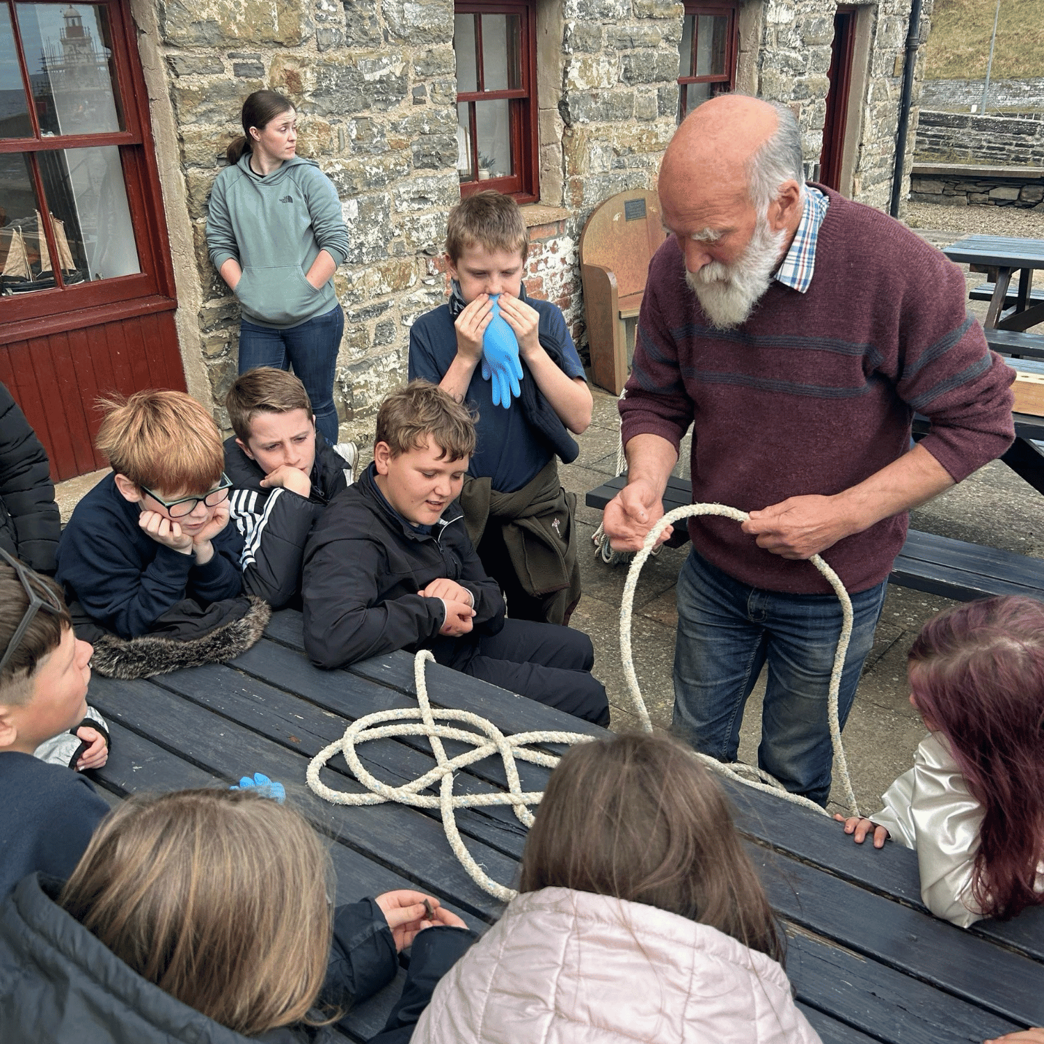 A man showing a group of 7 school pupils how to tie a knot in a large length of fishing rope.