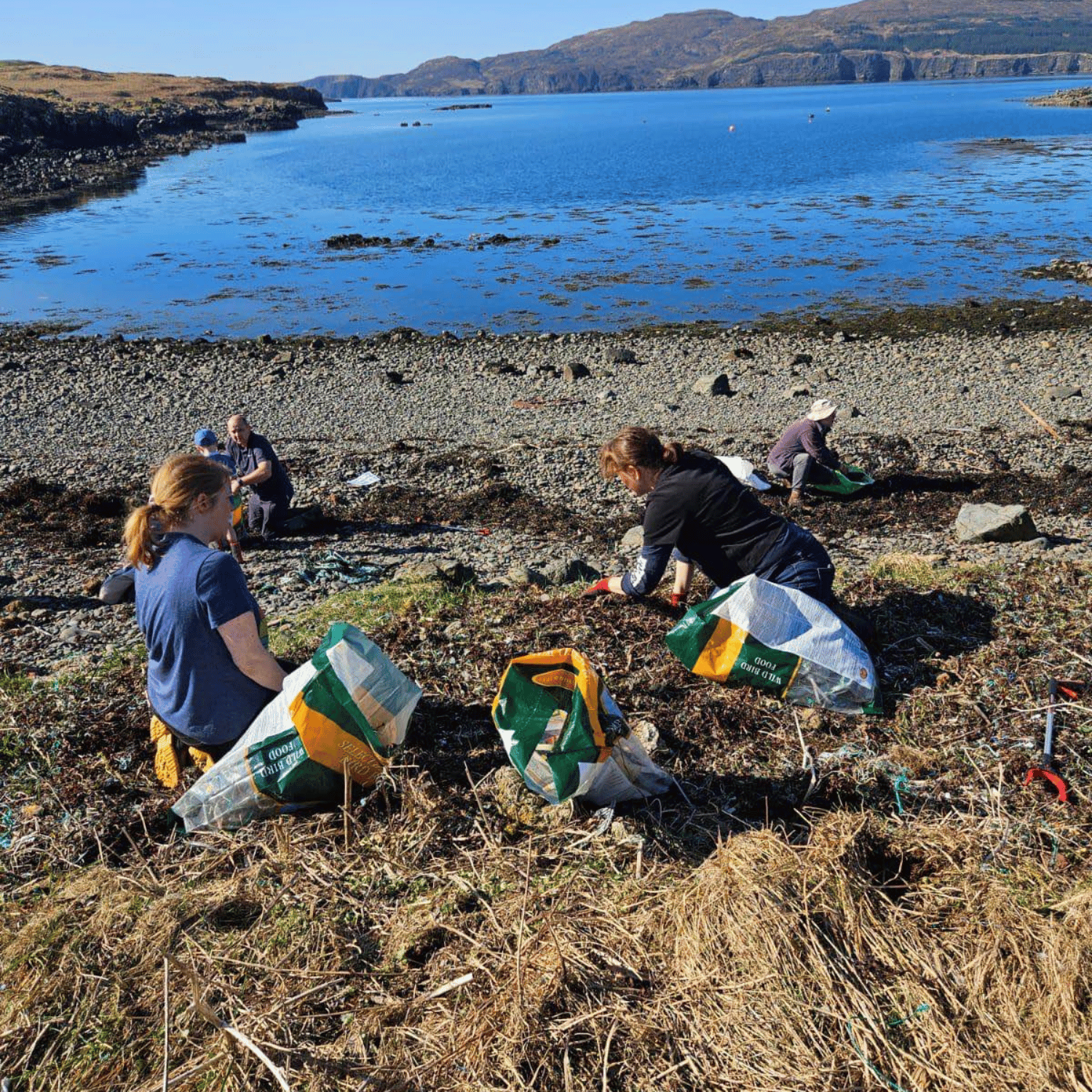 Two women picking litter from the banks of a loch. They are crouched down in the foreground filling up bags and in the background is the loch.