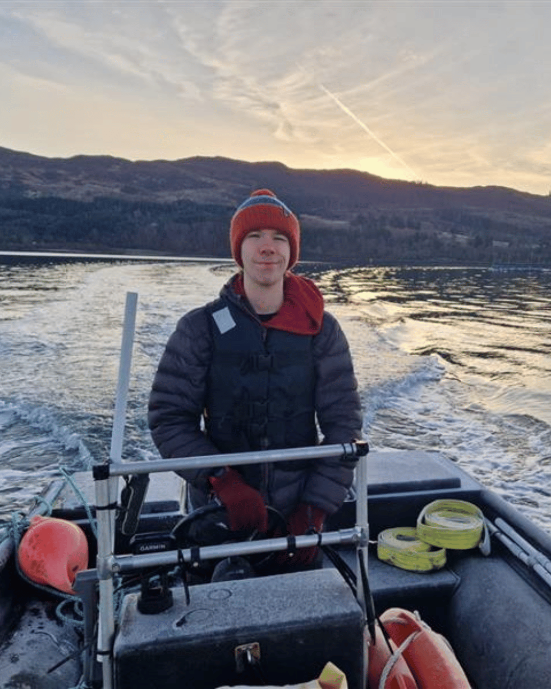 A teenage boy with a red hat and black jacket on driving a boat in a loch whilst the sun is setting behind him.