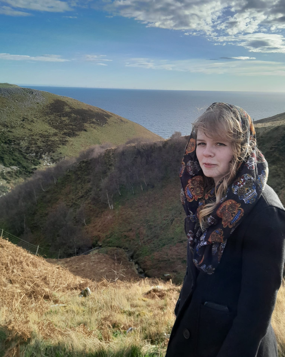 A teenage girl with blonde hair, a brown coat and purple scarf standing in a landscape of hills with the sea behind her.
