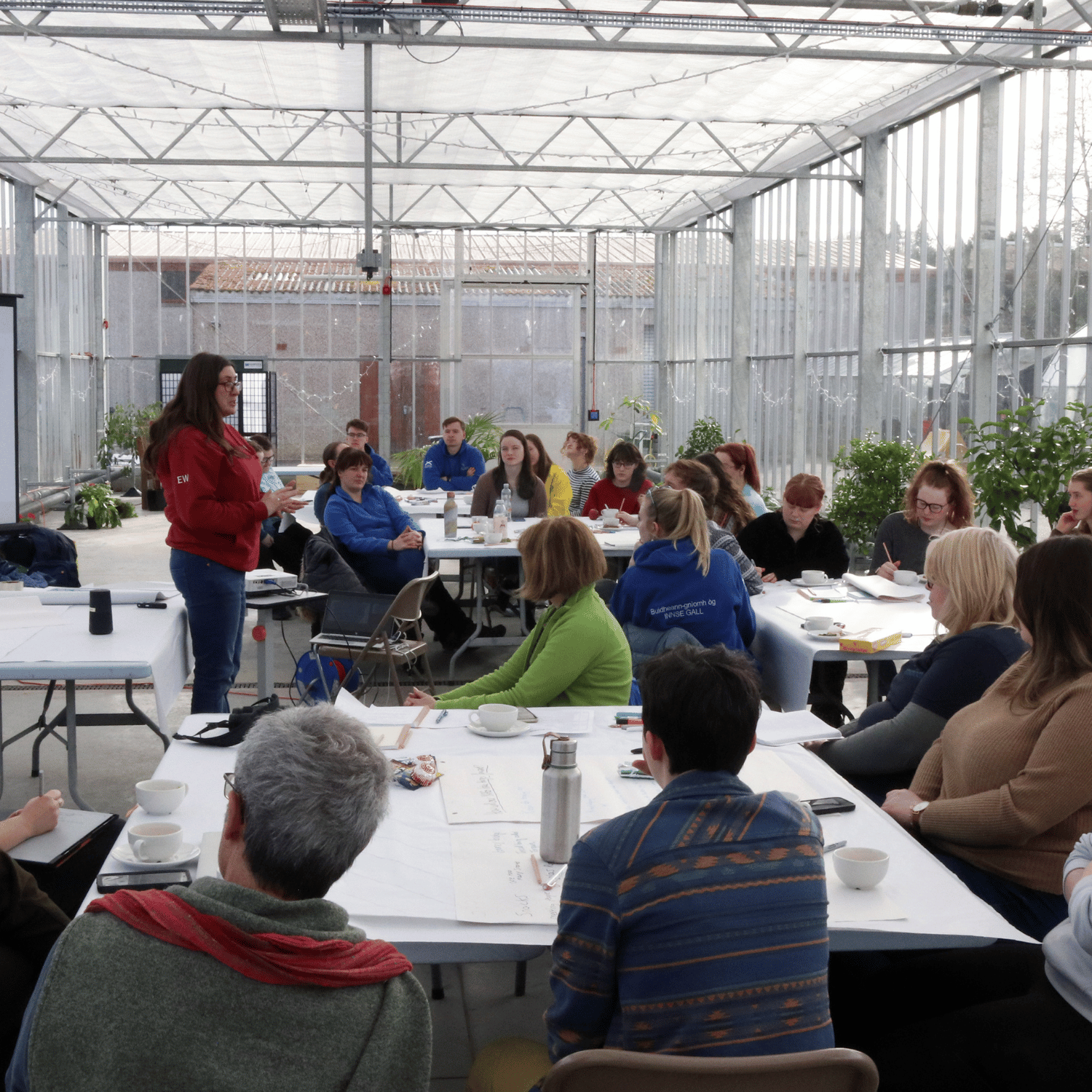 A room of young people watching a women in a red jumper and jeans give a presentation. Hosted in a greenhouse