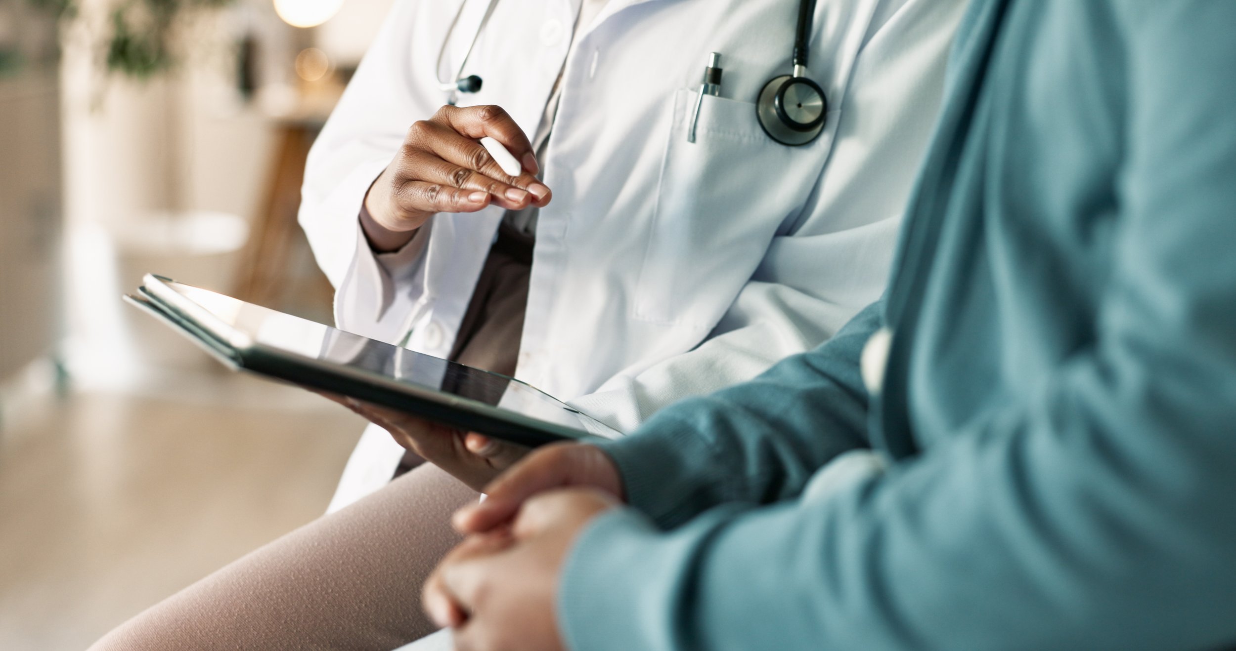 Healthcare professional in a white coat with a stethoscope looking at a tablet, sitting next to another person.