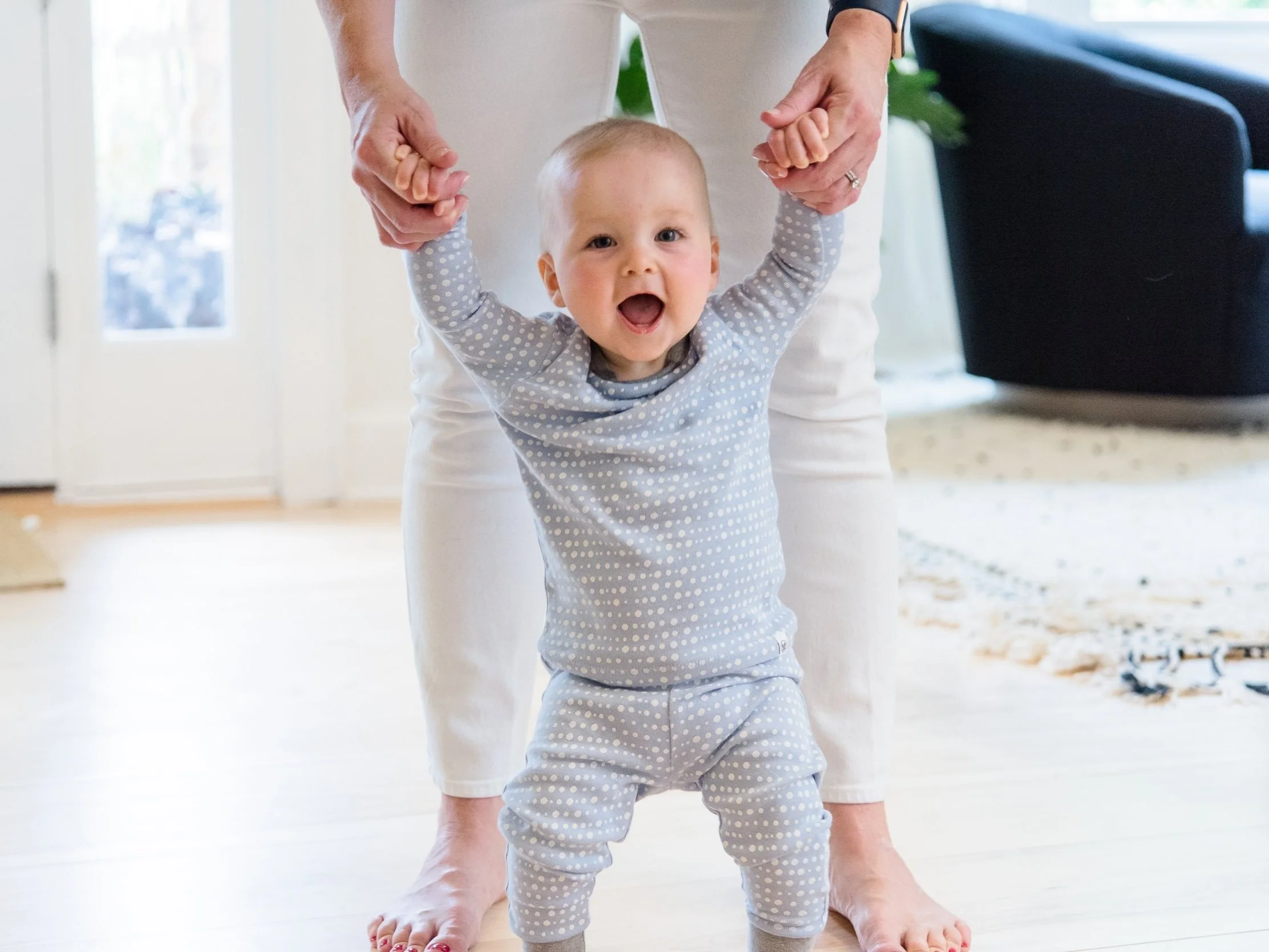 A happy baby standing between two adults supported by holding the adults' hands.The baby is smiling.