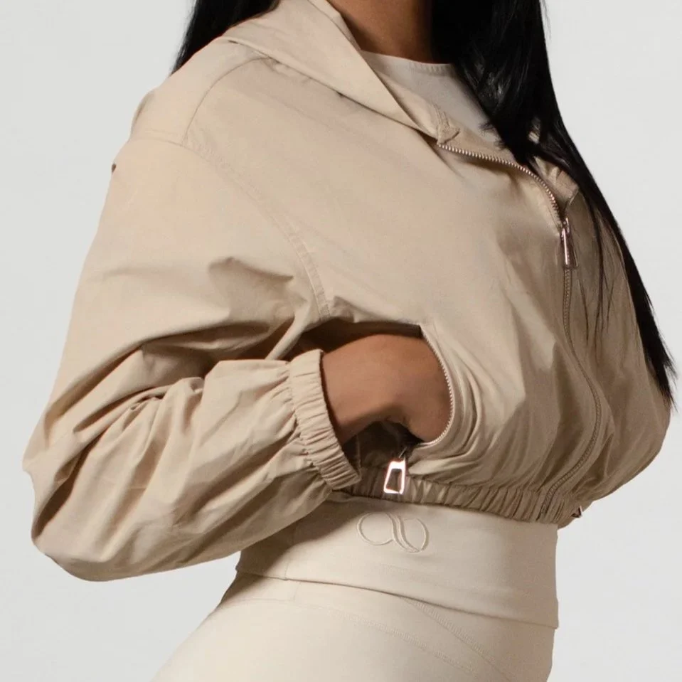 A woman wearing a beige cropped windbreaker with zippered pockets and elastic cuffs, standing against a plain background.